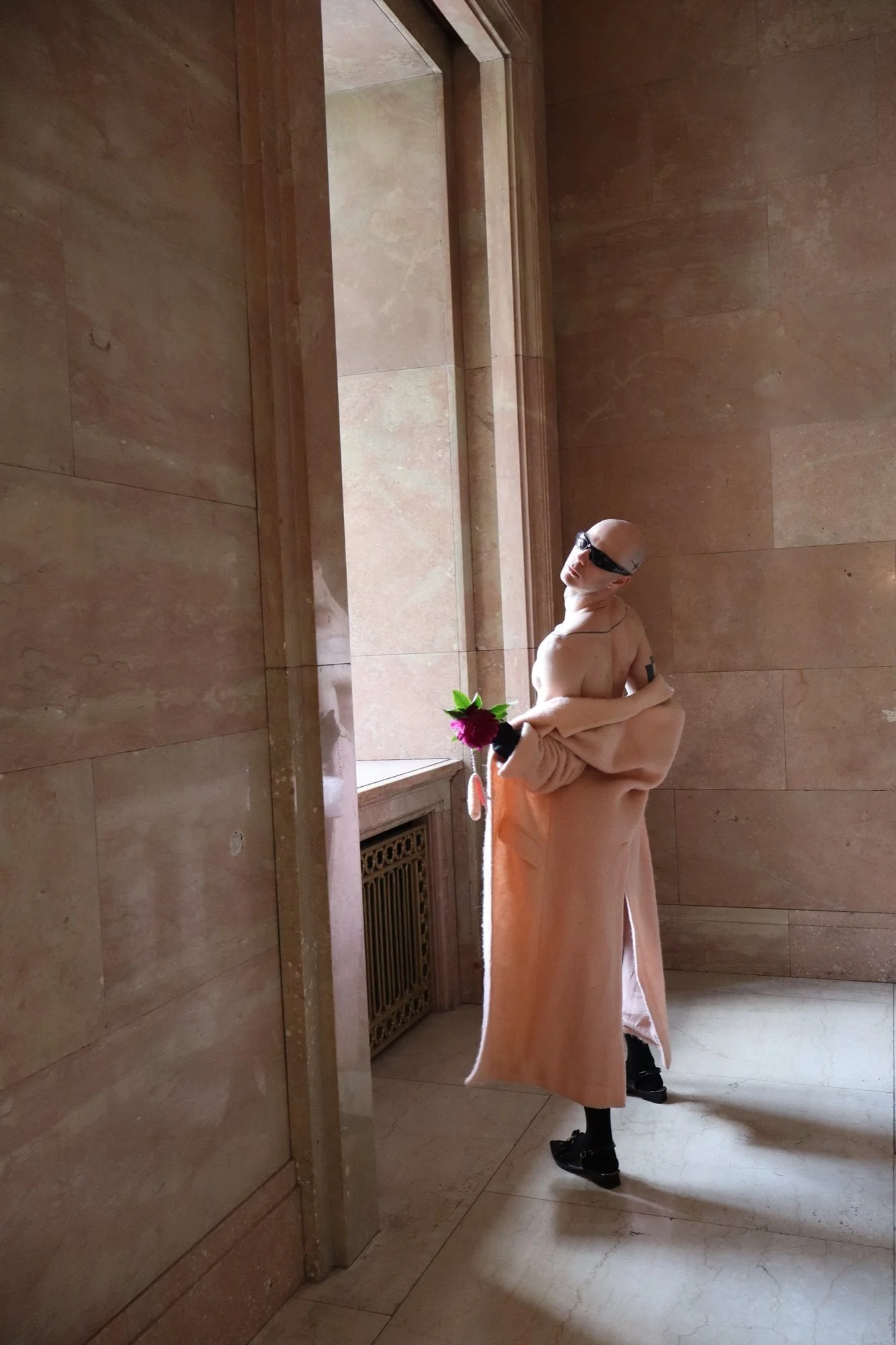 A person with a shaved head wearing sunglasses, a beige bathrobe, and black shoes, standing by a window with bare marble walls and floor, holding a pink flower.