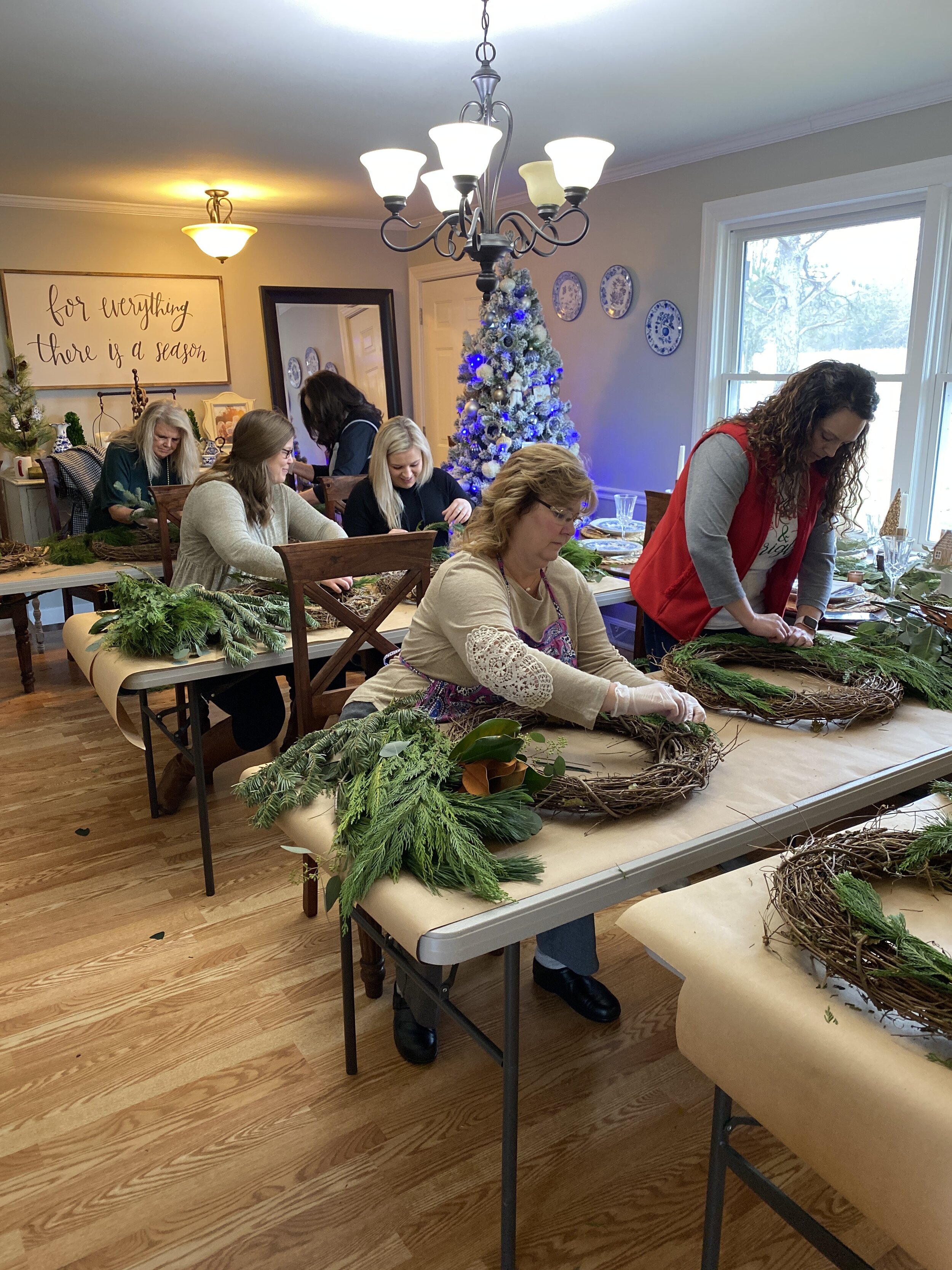 Group of women crafting Christmas wreaths in a decorated room with a Christmas tree and a "For everything there is a season" sign.