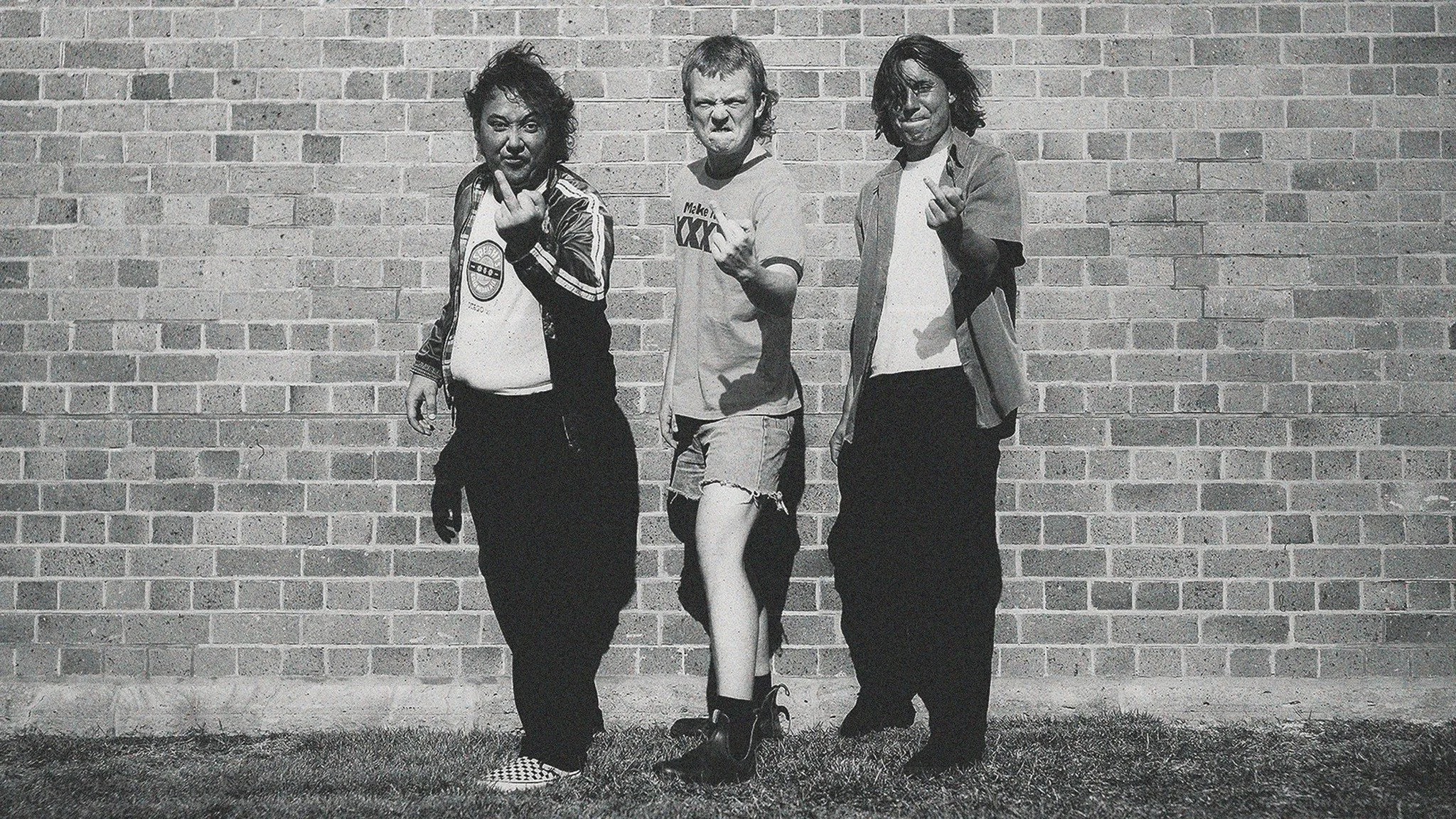 Three individuals posing confidently against a brick wall, with two pointing their fingers directly at the camera and one with a raised fist.