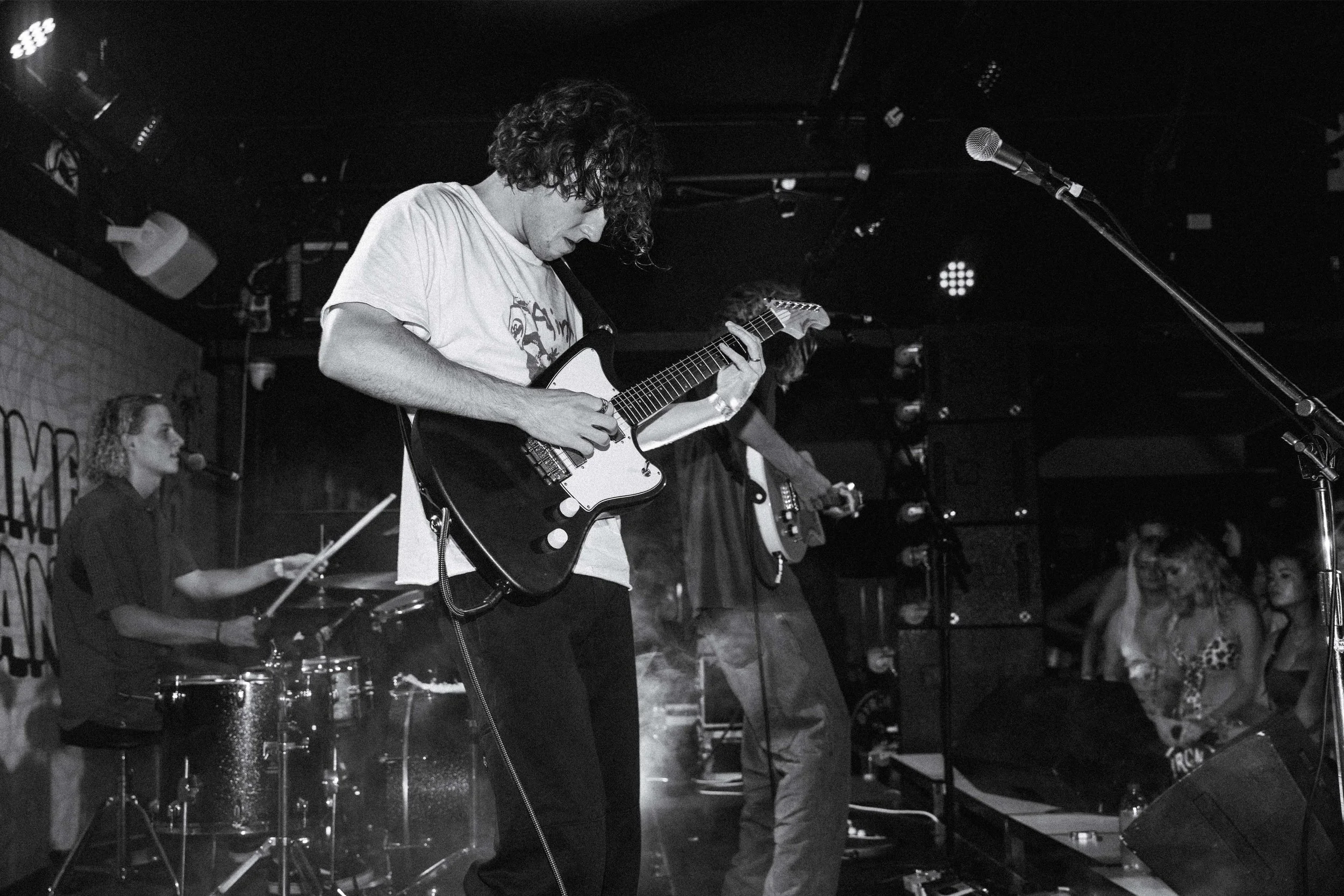 A live band performing on stage with a guitarist in the foreground, a drummer, and a bassist, in a black and white photo.