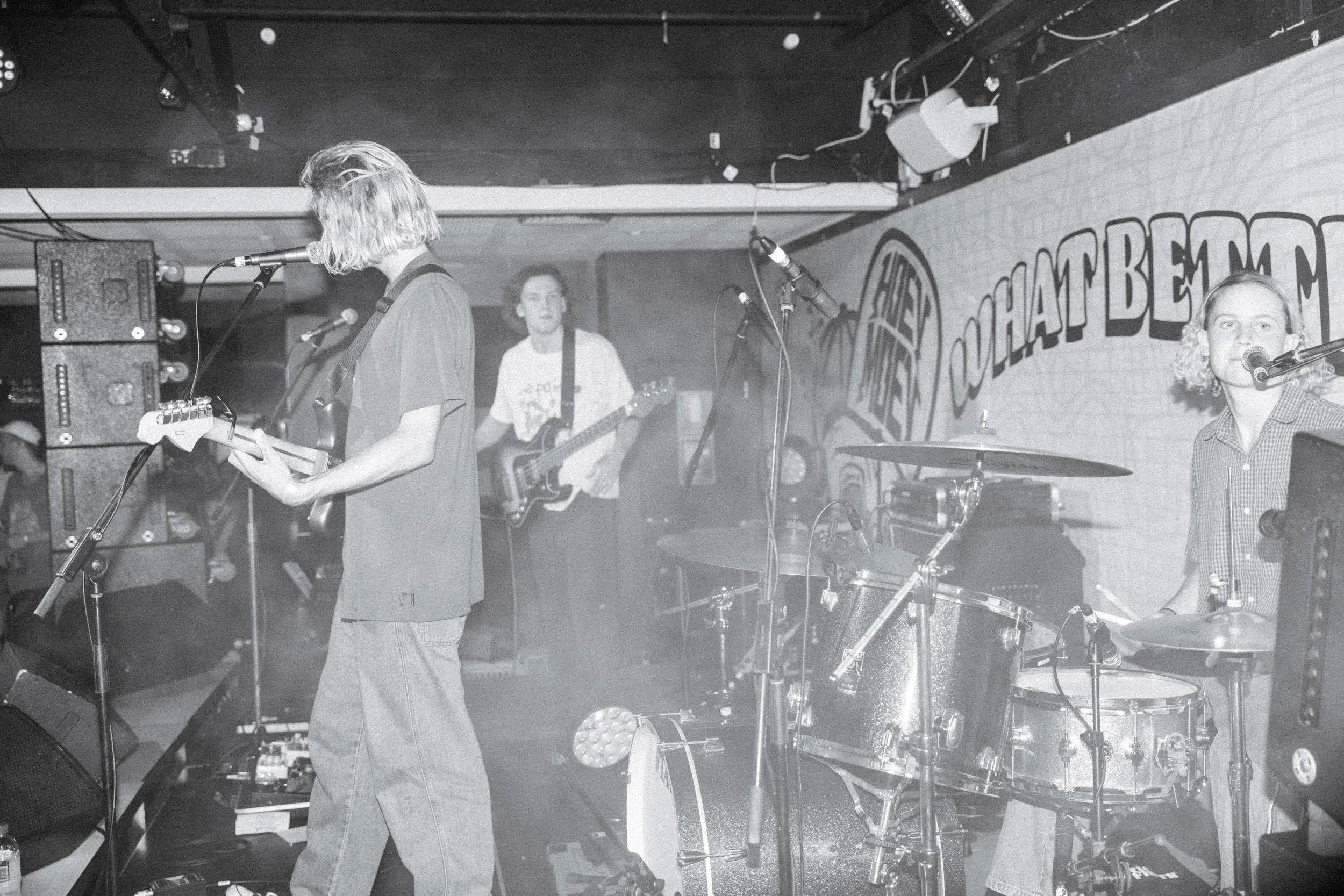 Three band members perform in a small venue with a graffiti-style mural in the background. One is playing guitar and singing into a microphone, another is playing bass guitar, and the third is playing drums.