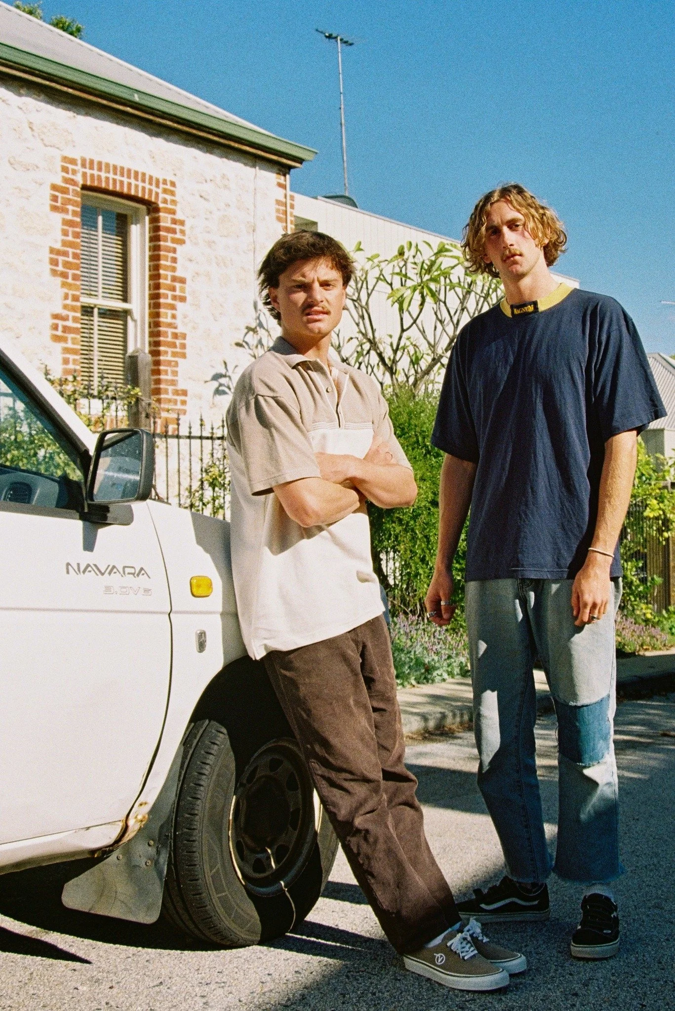 Two young men standing next to a white car on a street in front of a house with a brick and stone exterior, green window frames, and a garden. One man is leaning against the car with arms crossed, and the other is standing beside him.
