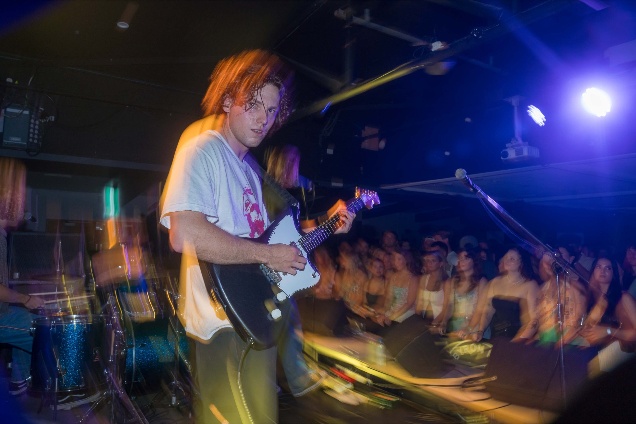 A young male musician playing an electric guitar on stage during a live concert with a crowd watching, colorful stage lighting, and a drum set visible in the background.