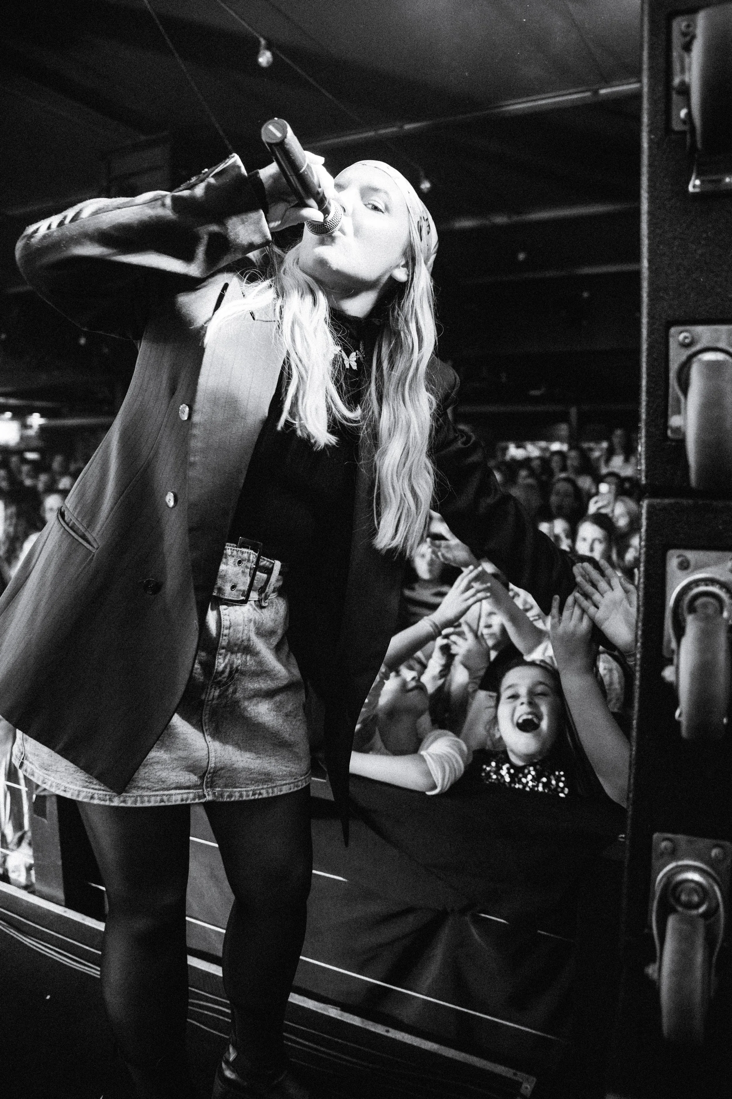 A female singer with long hair singing into a microphone on stage while an enthusiastic crowd reaches out to her.