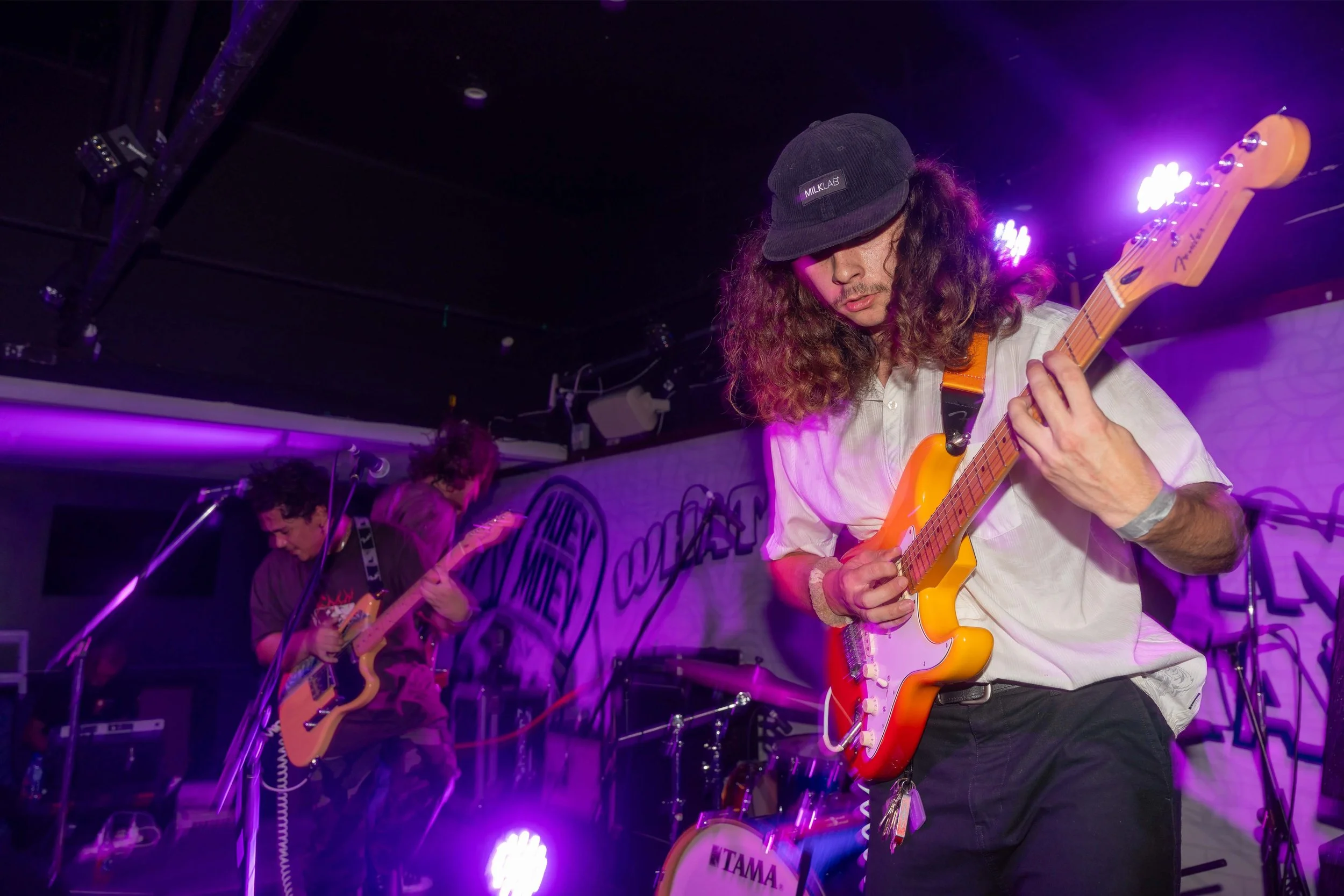 A band performing on stage under purple lighting, featuring musicians playing electric guitars.