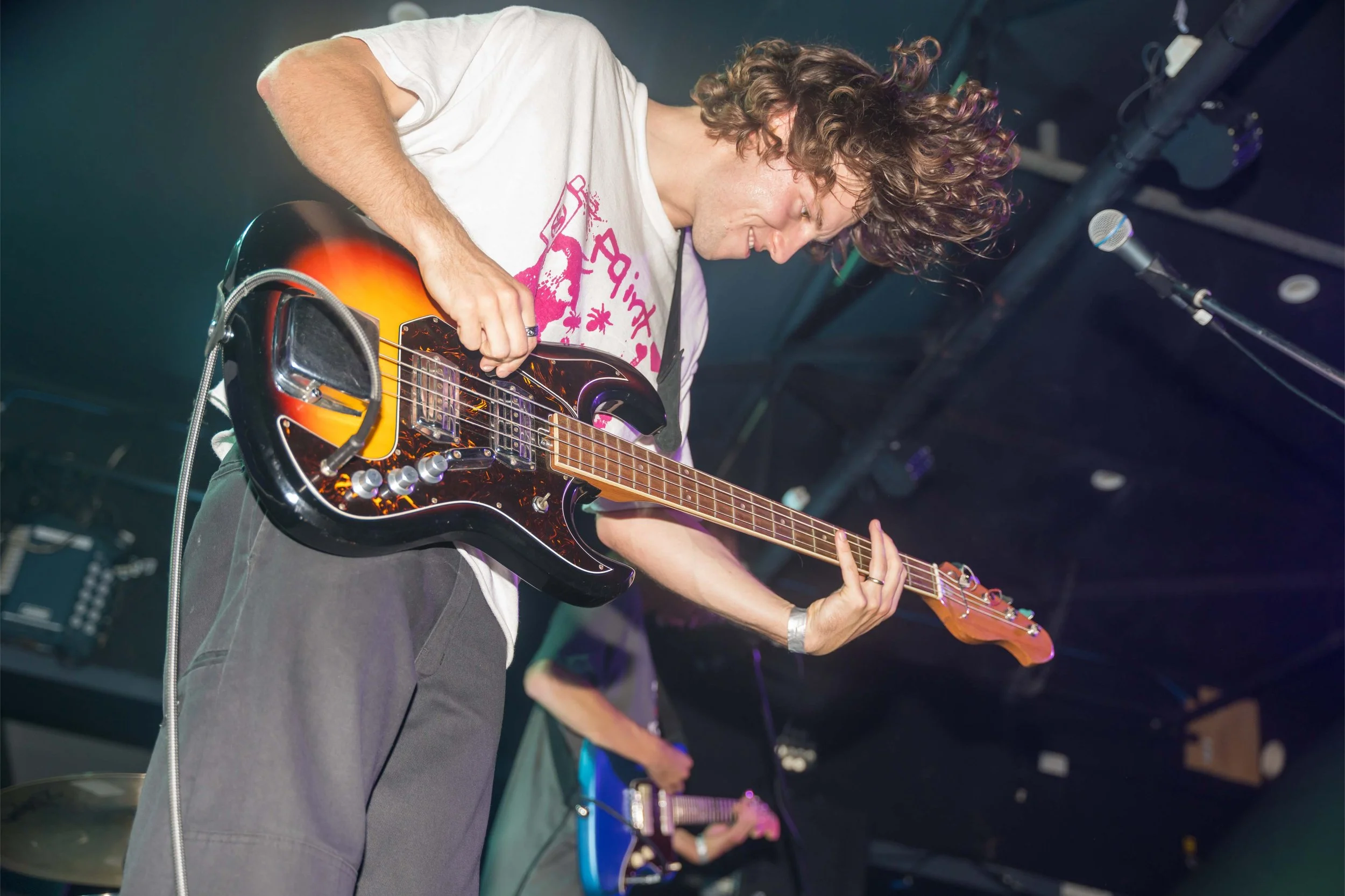 Musician playing electric guitar on stage with a mic in front, stage lighting in the background.