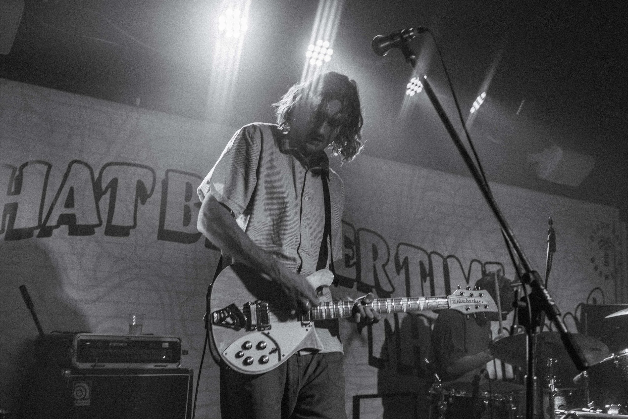 A musician playing an electric guitar on stage under bright lights, with a drum set nearby and a microphone stand in front.