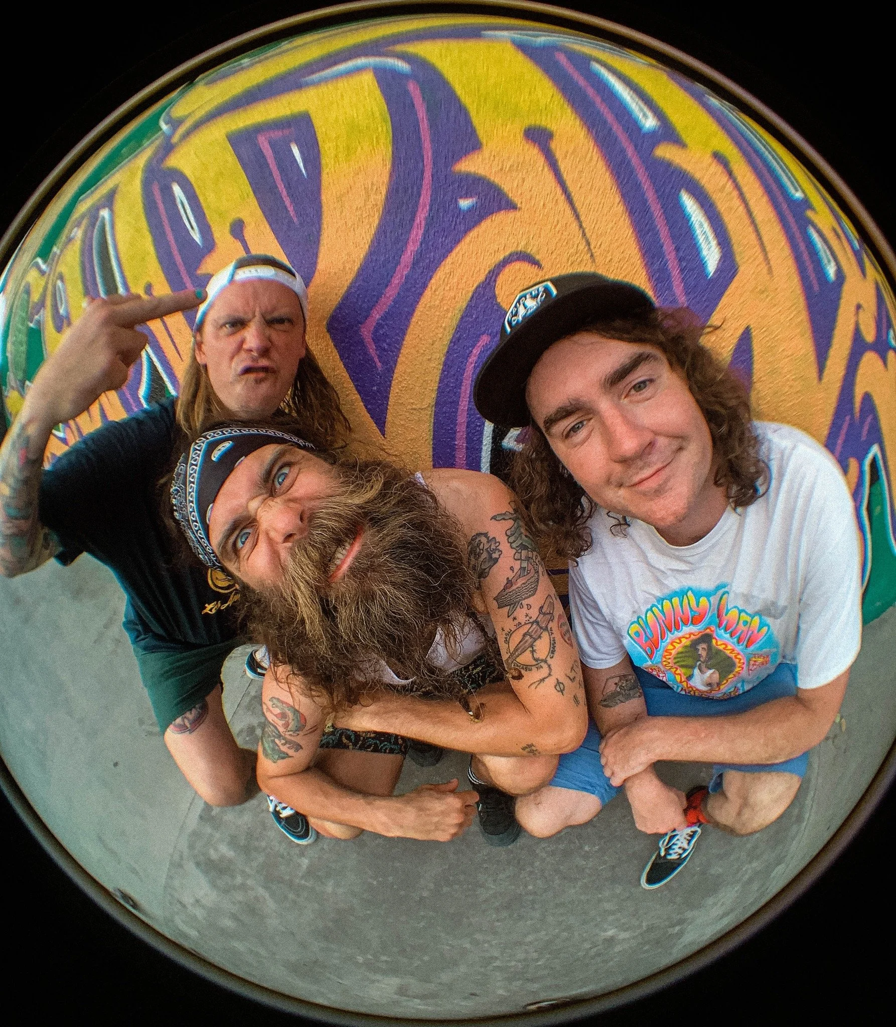 Three young men with tattoos and casual clothing crouching in front of a colorful, graffiti-style wall, taken with a fisheye lens.