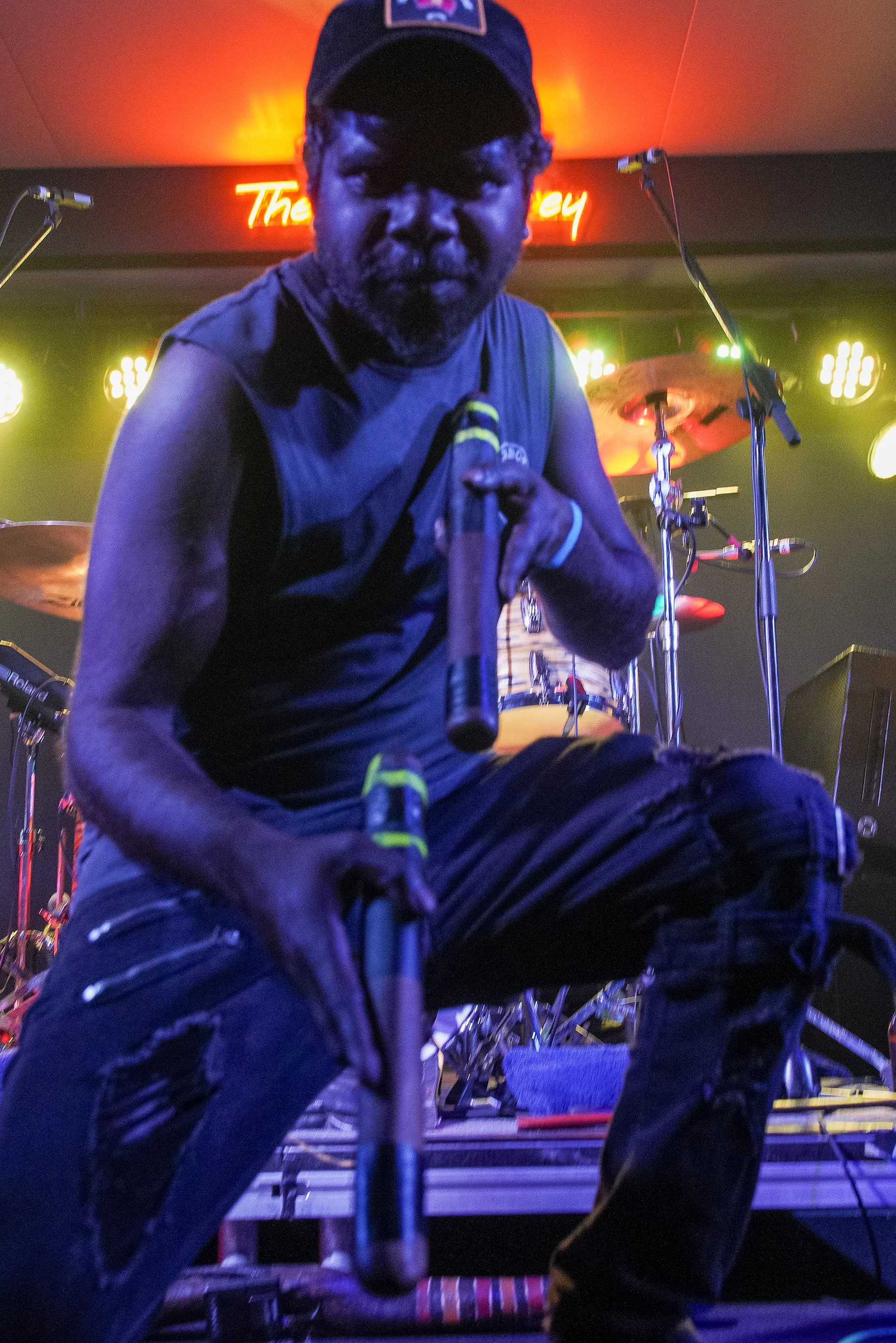 A man holding a microphone, kneeling on stage with musical equipment and drum set in the background, neon sign and stage lights in the background.