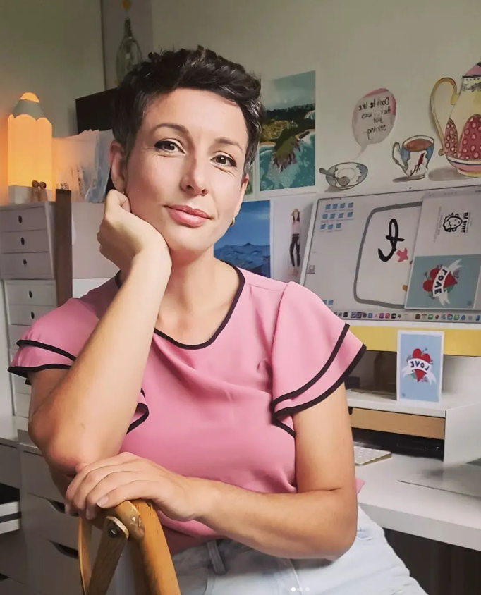 Woman with short dark hair sitting at a desk in a colorful, creative workspace, wearing a pink top with black trim, resting her chin on her hand and smiling at the camera.