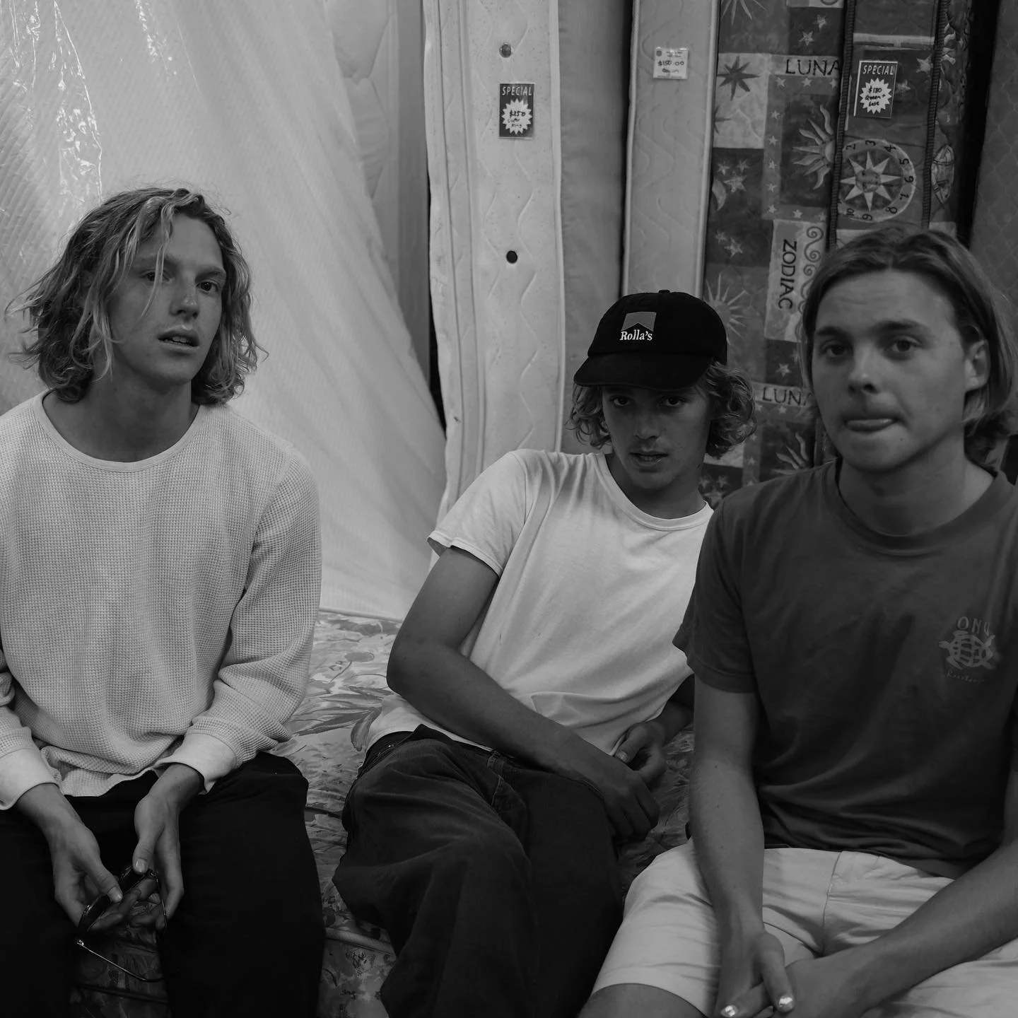 Three young men sitting on a bed in a room with patterned curtains and various signs, two of them are alternatingly looking at the camera, while the third is looking slightly intently to the side, with the man on the right sticking out his tongue.
