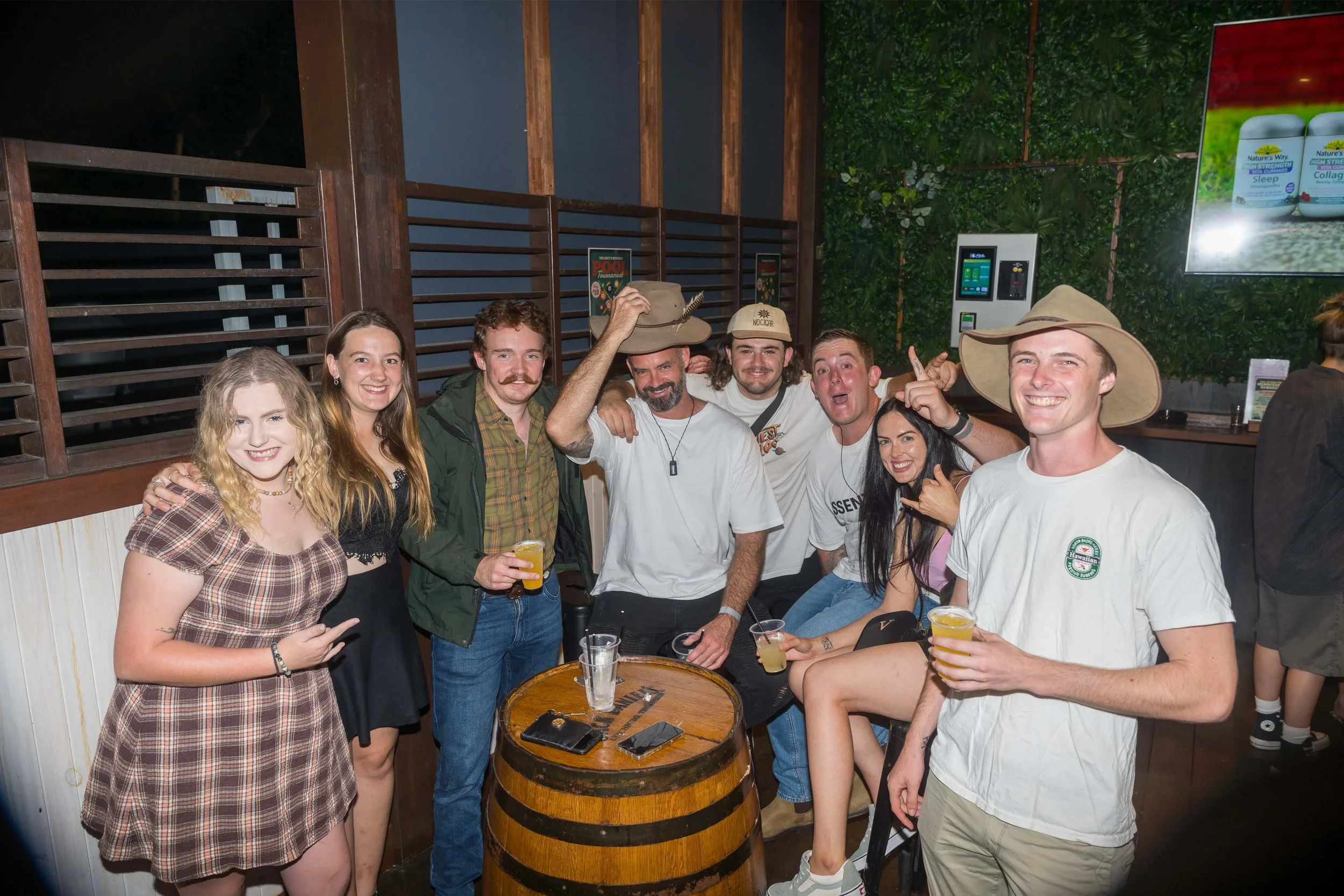 Group of friends at a bar celebrating, some wearing cowboy hats, smiling, with drinks in hand, standing around a wooden barrel table in a rustic-style venue.