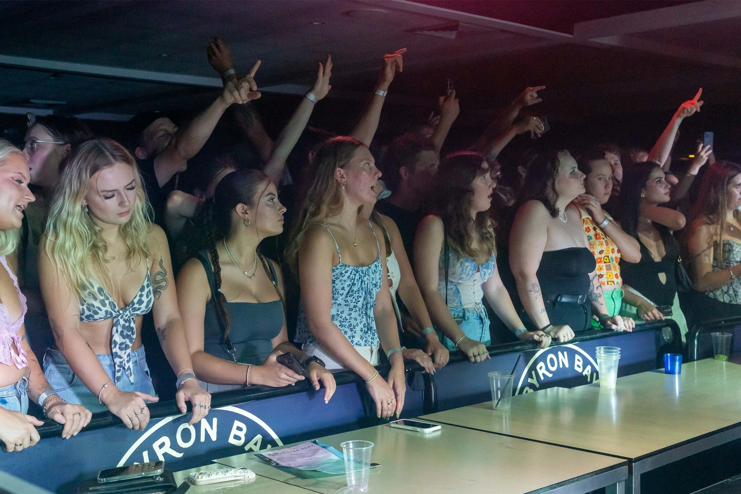 Group of young women at a concert or event standing behind a barrier, some raising their hands, with drinks on the table in front.