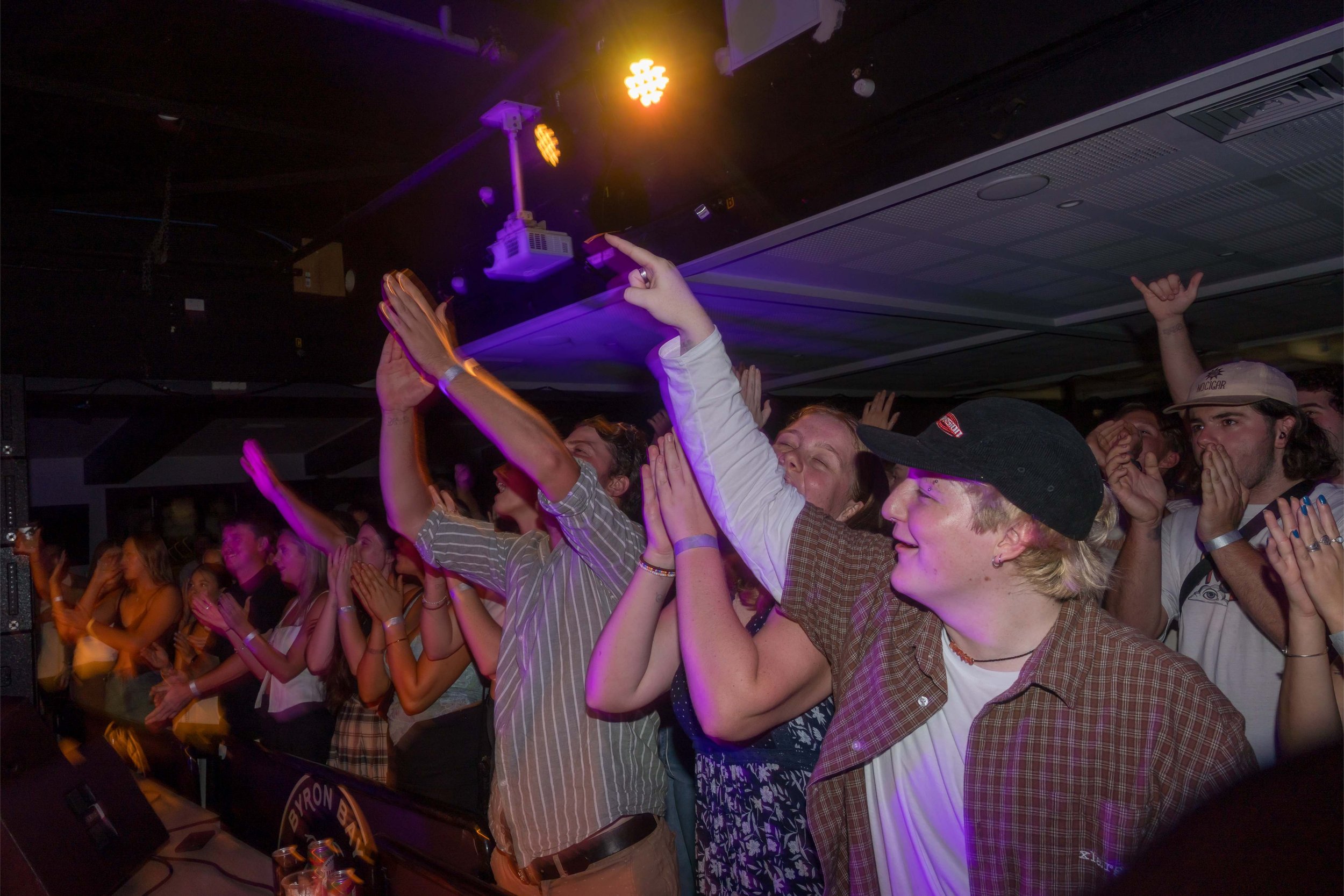 A lively crowd of young people at a concert or music event, cheering and enjoying the performance under colorful stage lights.