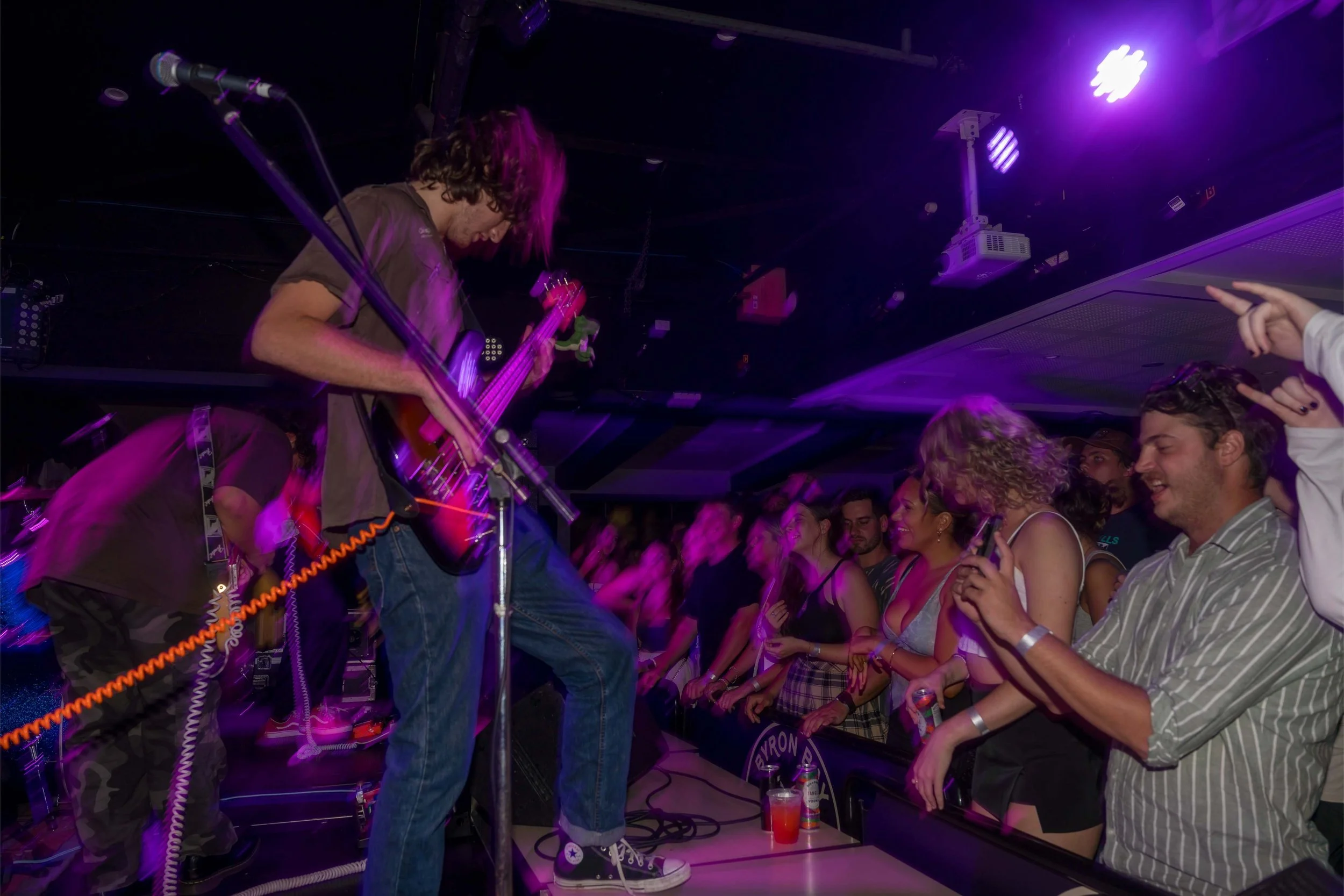 Musicians performing on stage with an audience enjoying the concert, vibrant purple stage lights, and a man in striped shirt holding a smartphone and smiling.