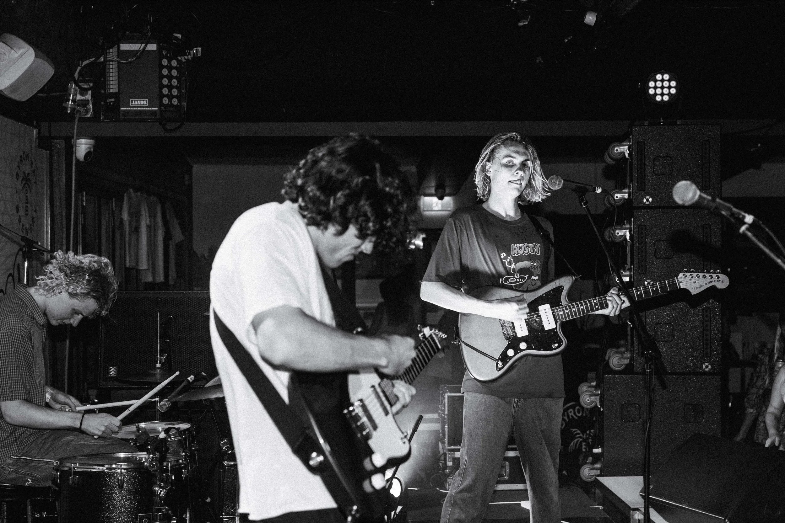 Three musicians performing on stage: a drummer in the background, a guitarist in the foreground, and a female vocalist playing an electric guitar, all in black and white.