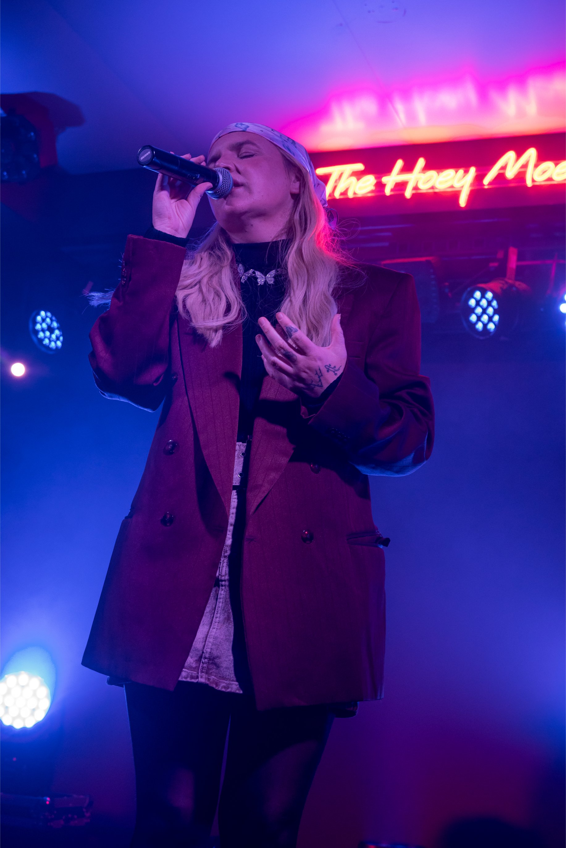 A woman with long blonde hair, wearing a bandana, maroon blazer, and black tights, is singing into a microphone on stage with a neon sign in the background that reads 'The Honey Moon.' She has tattoos on her fingers and a hand on her chest, closed eyes, and appears emotionally engaged in her performance.