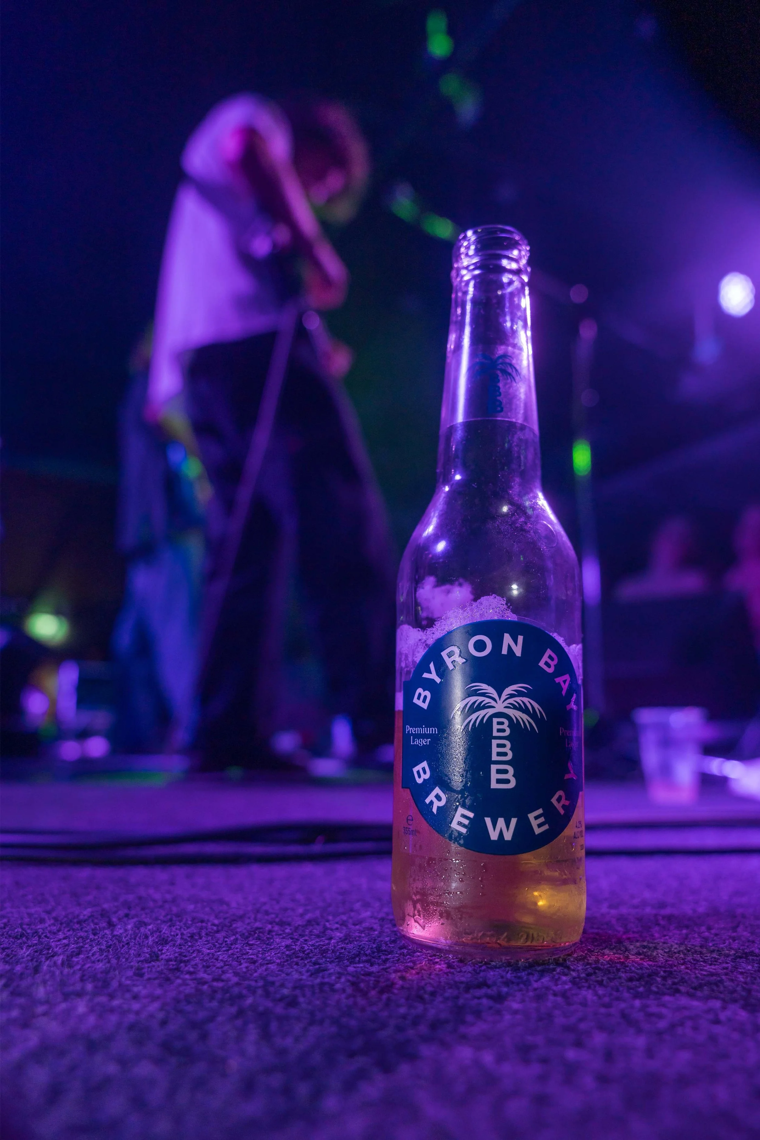 A beer bottle from Byron Bay Brewery with a partially consumed beverage on a textured surface, with a blurred dancer performing in the background under vibrant purple and green lighting.