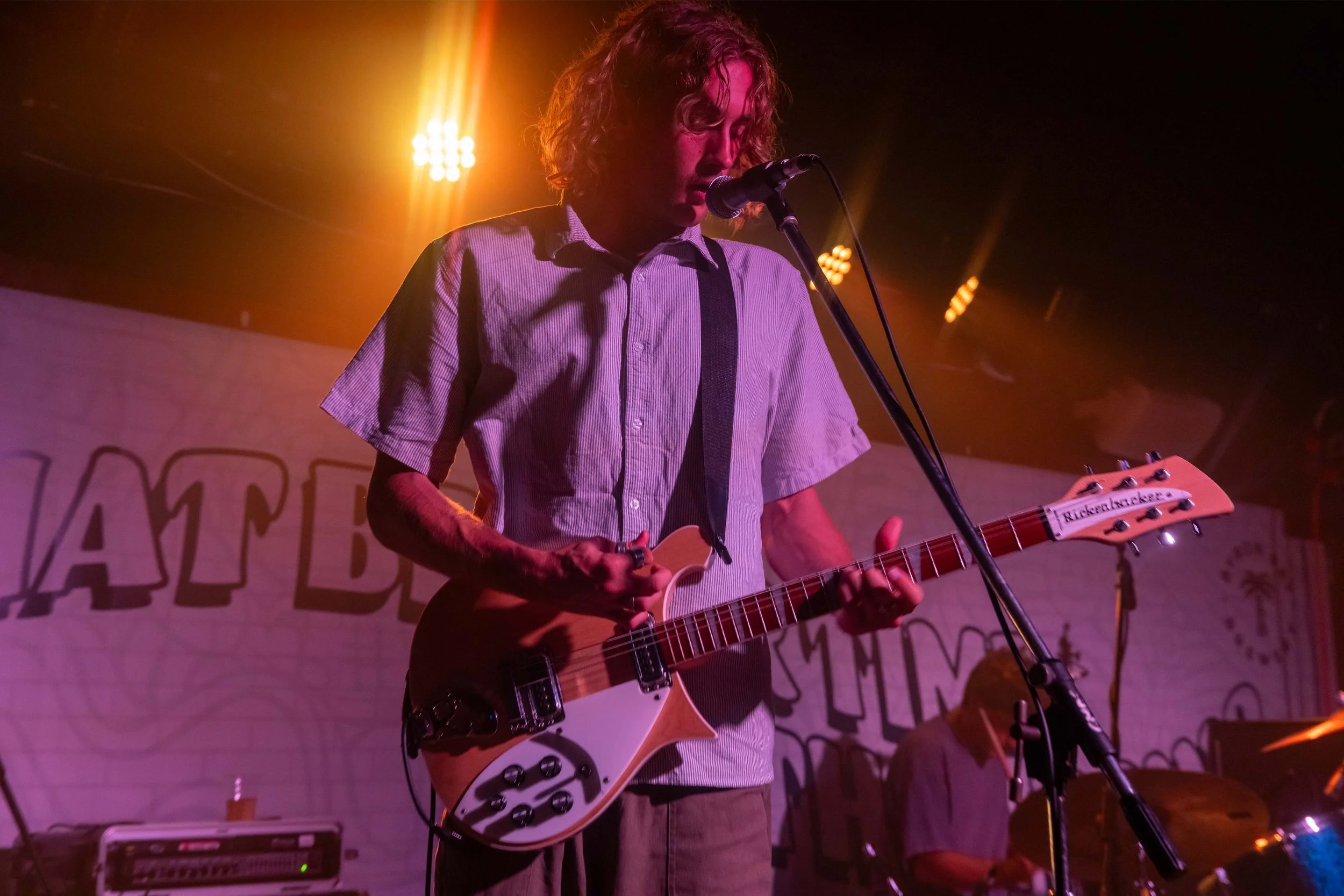 A man with curly hair playing an electric guitar on stage with a microphone, illuminated by stage lights, with a background banner and another musician.