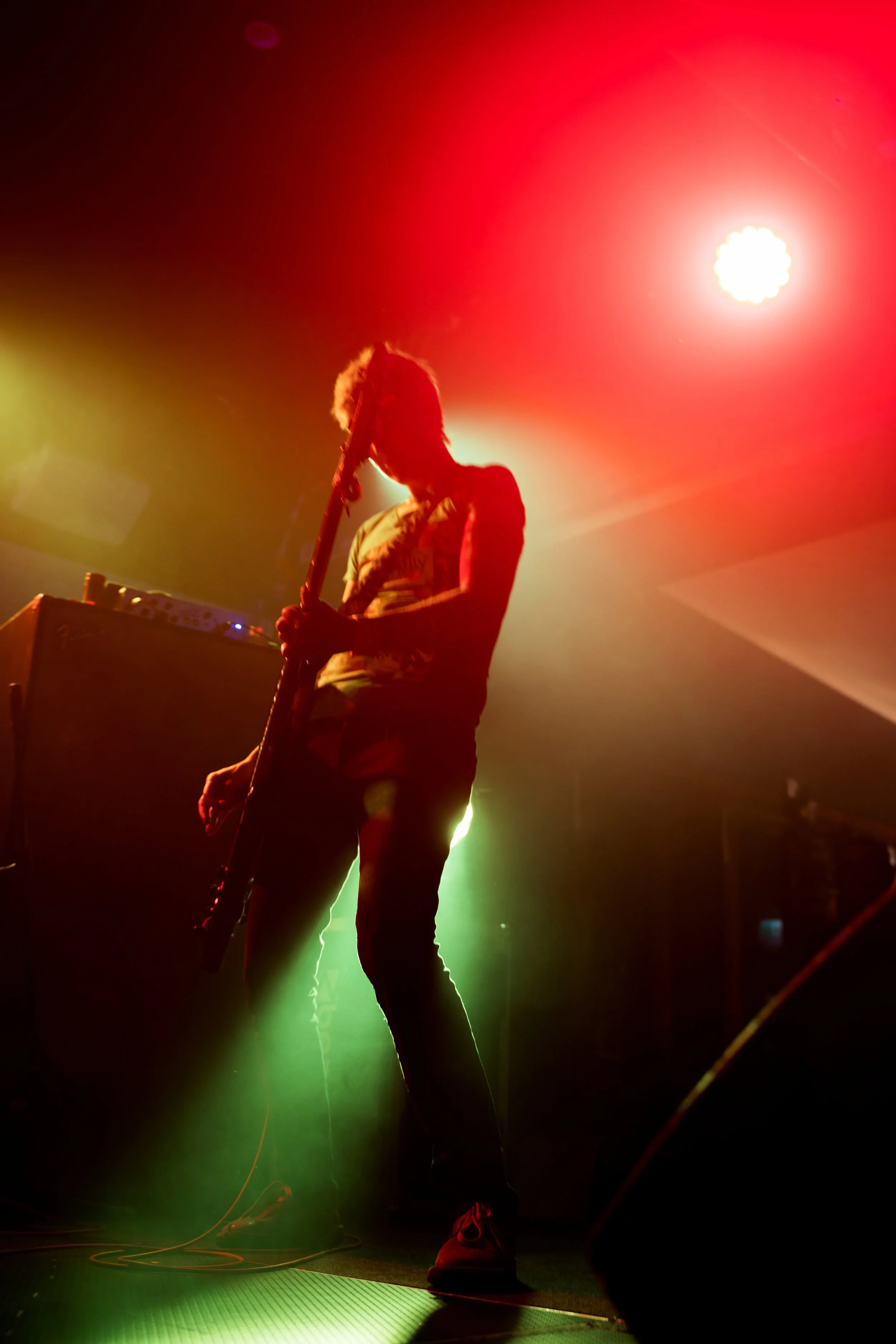 Musician playing an electric guitar on stage with colorful red, yellow, and green lights creating a vibrant atmosphere.