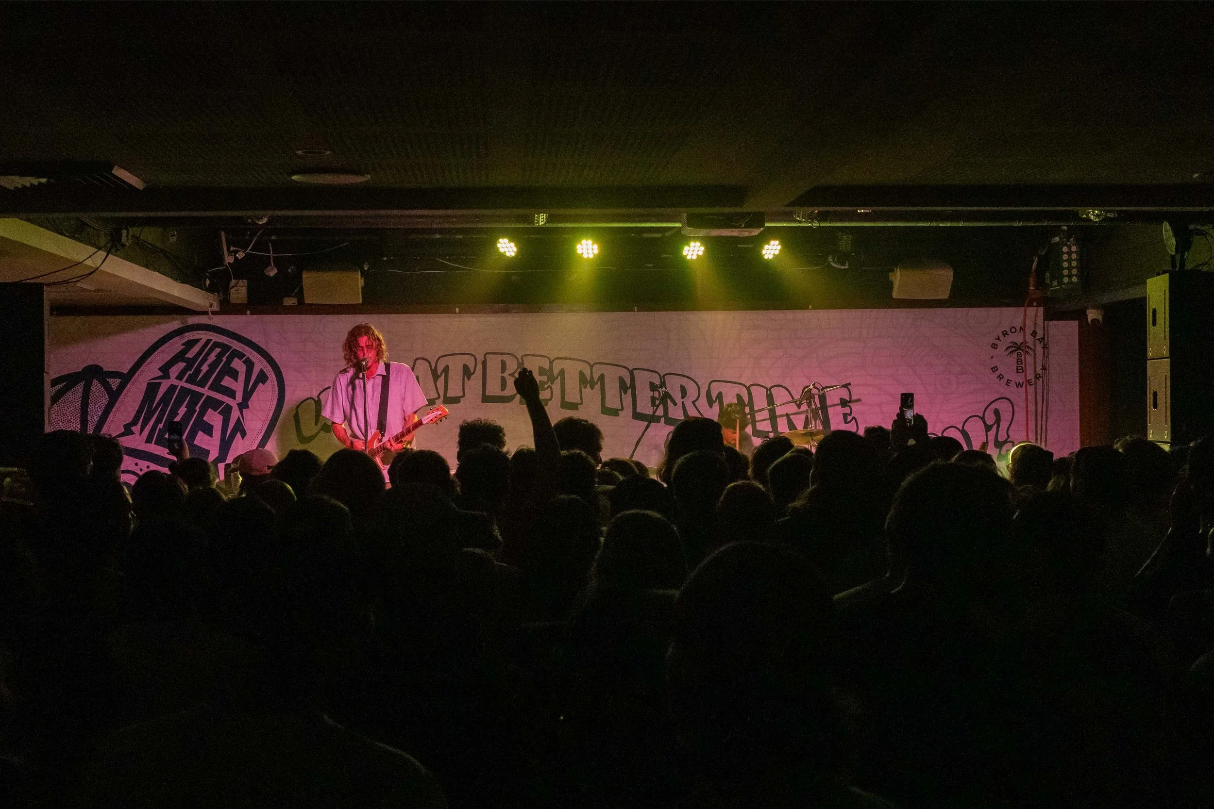 A musician with curly hair playing an electric guitar and singing into a microphone on stage during a live concert. The stage backdrop has colorful graffiti-style text that says, 'Get Better Together' and other words, with stage lights overhead.