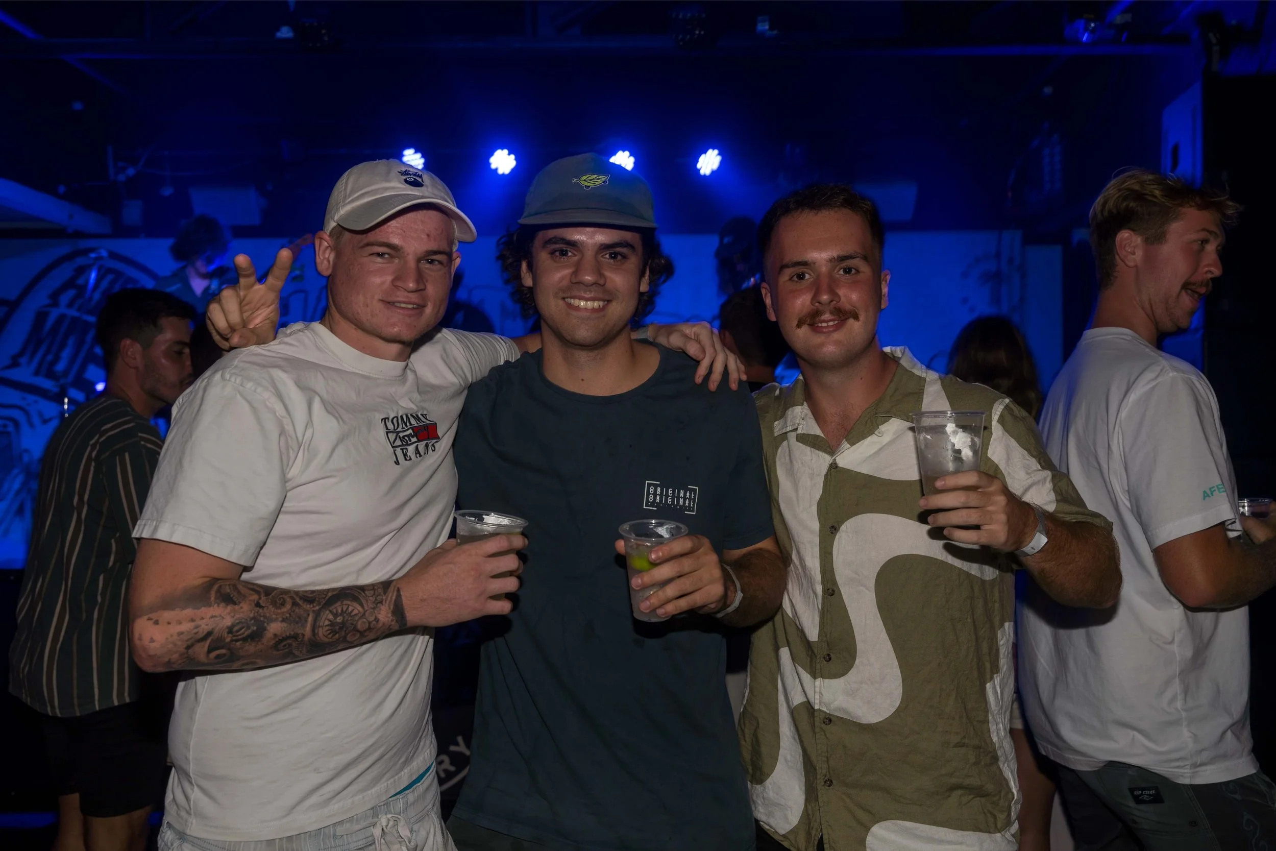 Three young men happily posing with drinks in a nightclub with blue lighting, some with tattoos and casual summer clothing.