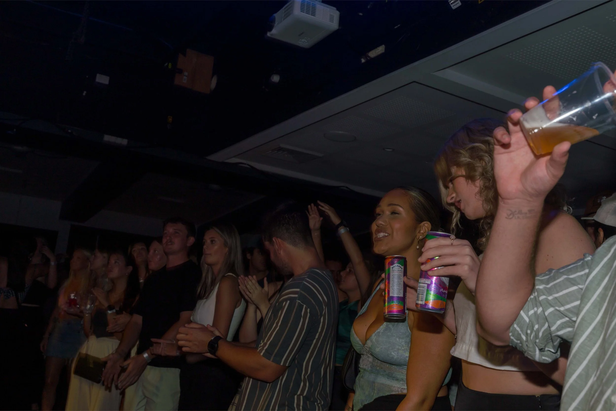 Crowd of people at a party or concert, some holding drinks, with a dark background and ceiling lights.
