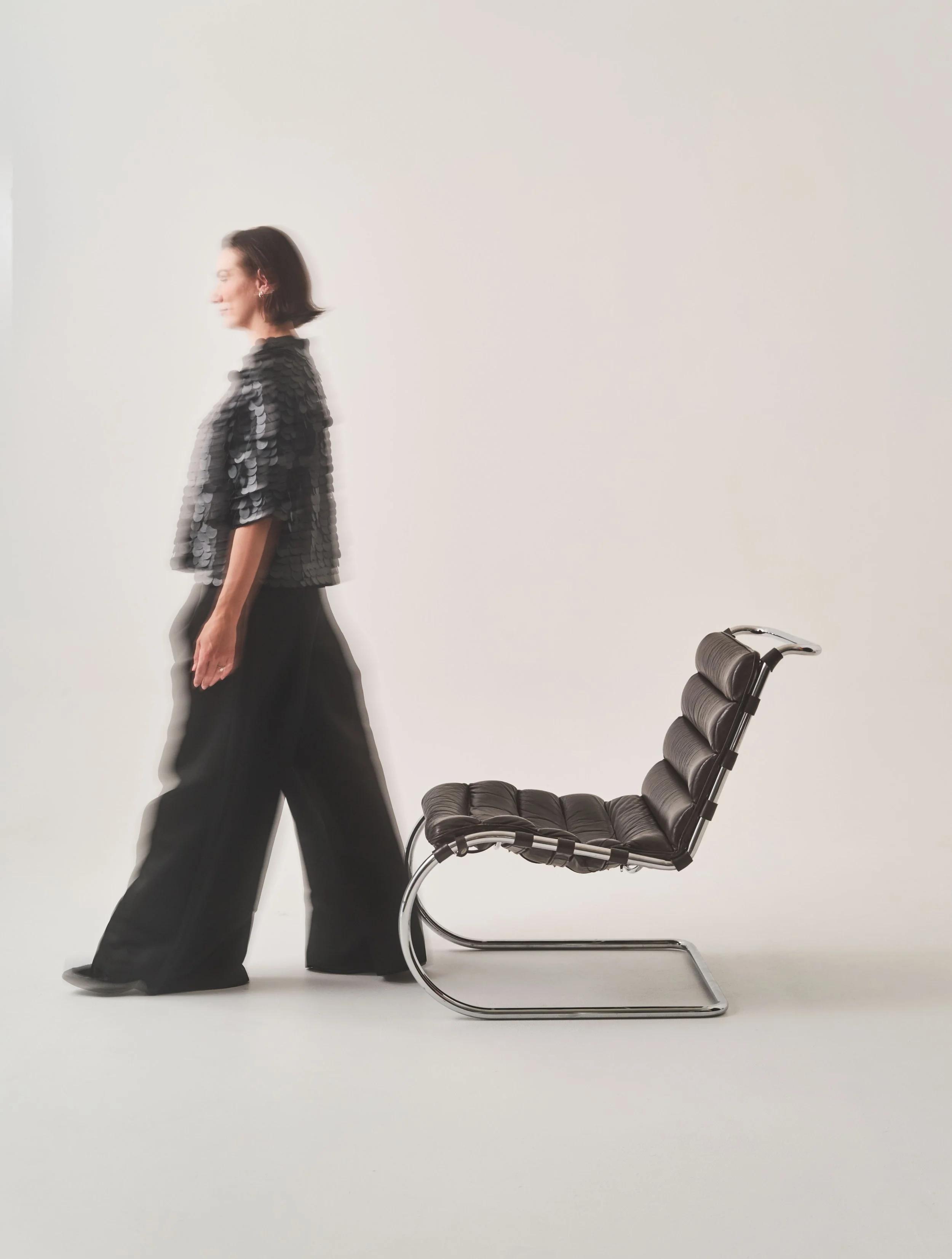 A woman with dark hair wearing a black sequined top and wide-legged black pants standing next to a black leather recliner chair on a white background.