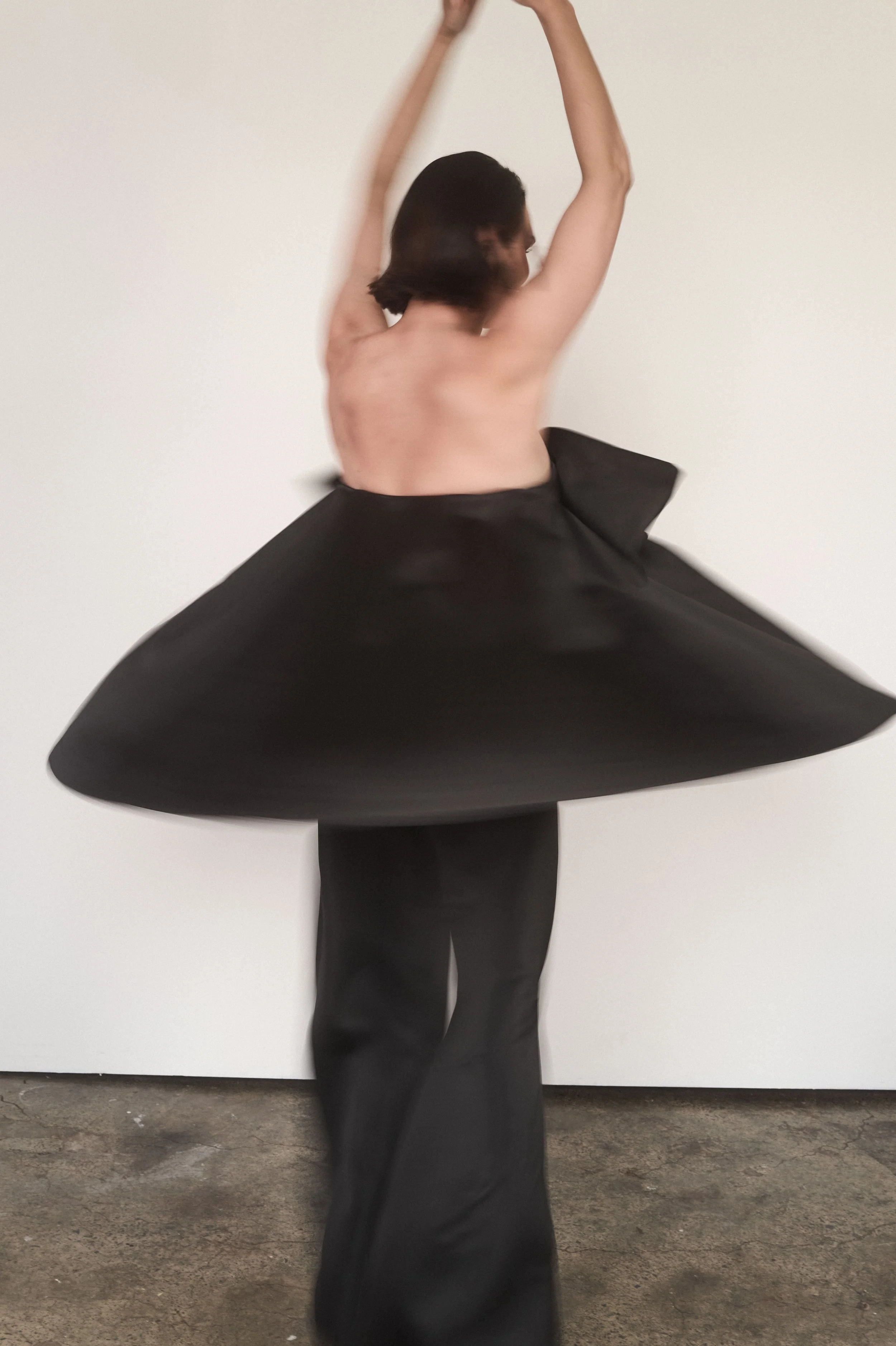 A person with short dark hair, seen from behind, wearing loose black pants and no top, dramatically posed against a plain white background. The photo is slightly blurred.