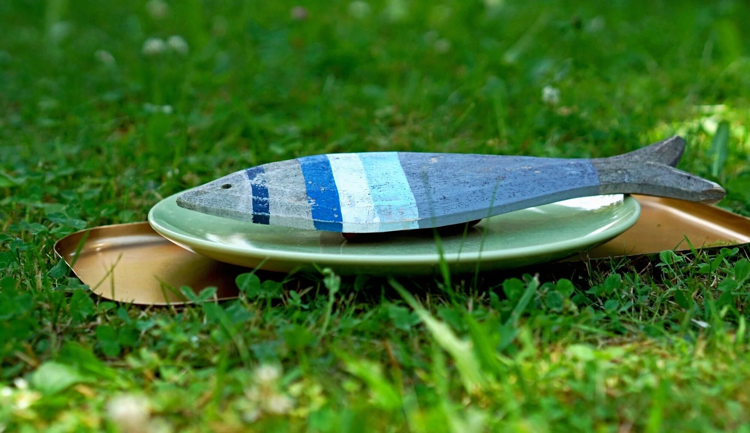 Ein auf einer grünen Wiese liegendes graues Holzfisch-Spielzeug mit blauen Streifen, auf einem grünen Teller, auf einem goldfarbenen Tablett.