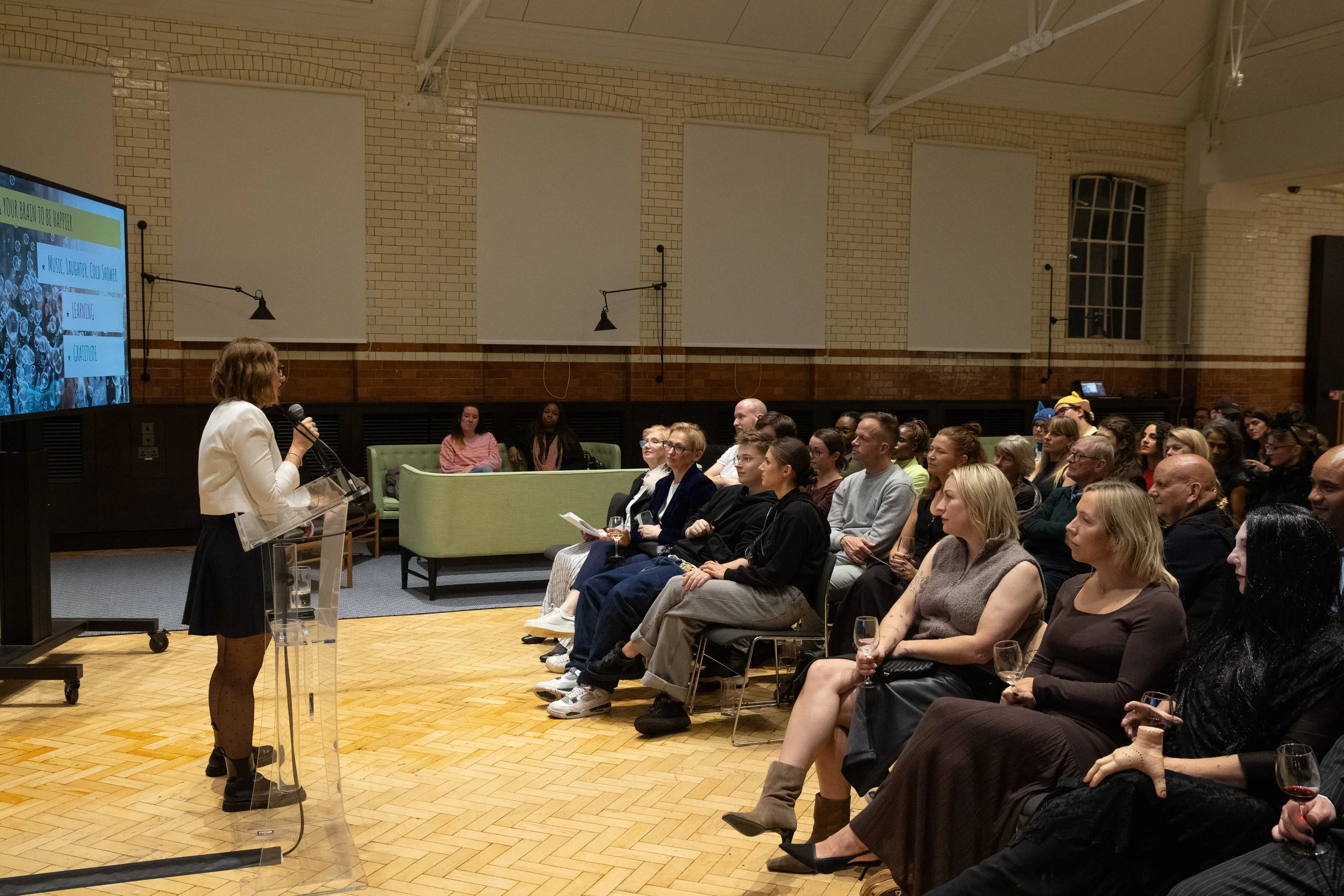 A woman with shoulder-length brown hair, glasses, and a light-colored blazer is sitting at a desk, smiling and looking at the camera, with a laptop in front of her.