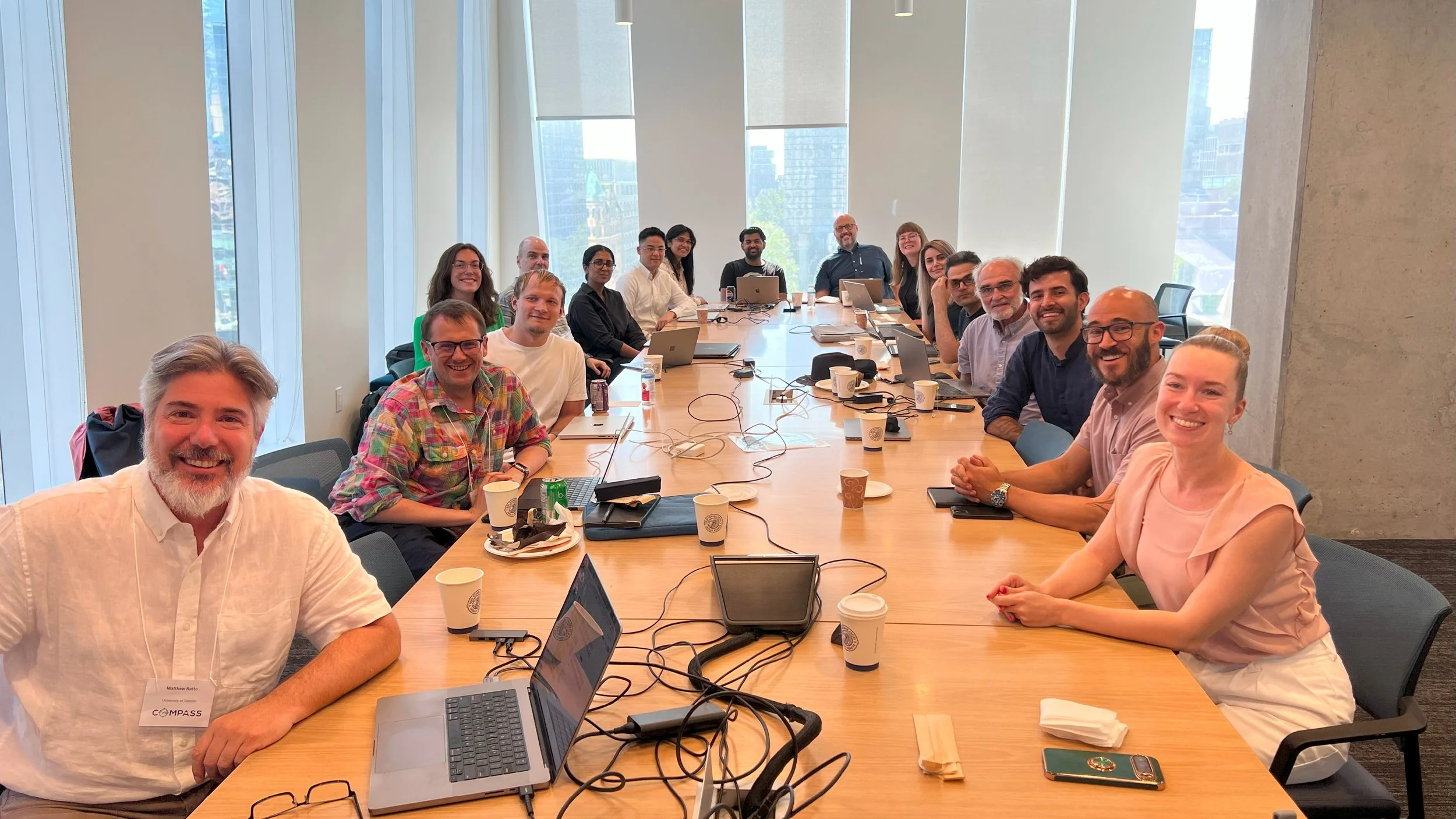 A group of smiling people sit around a long boardroom table covered in coffee cups, laptops, and cables.