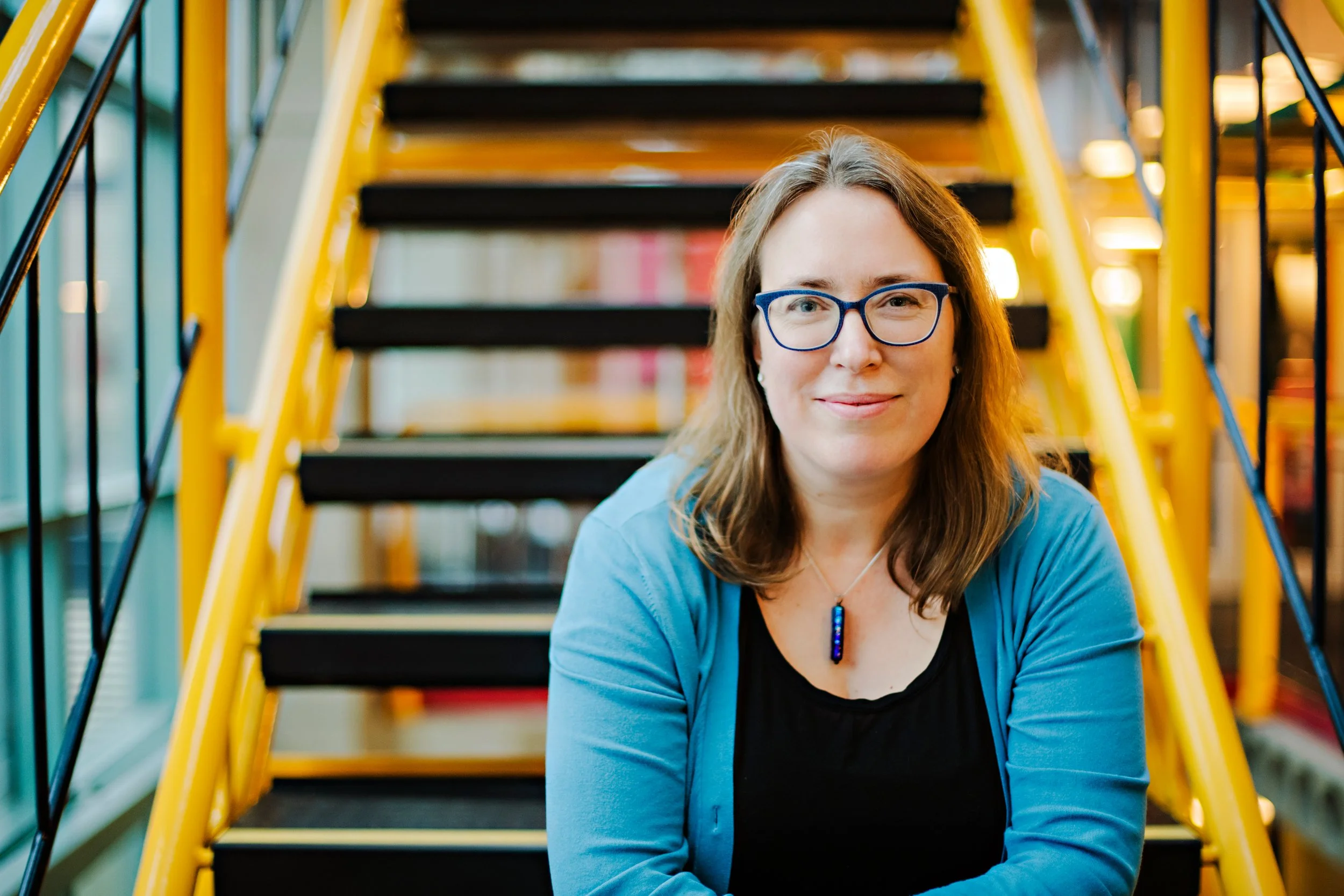 Kate Larson poses for a headshot wearing glasses as well as a green cardigan with a black top, seated in front of a flight of stairs