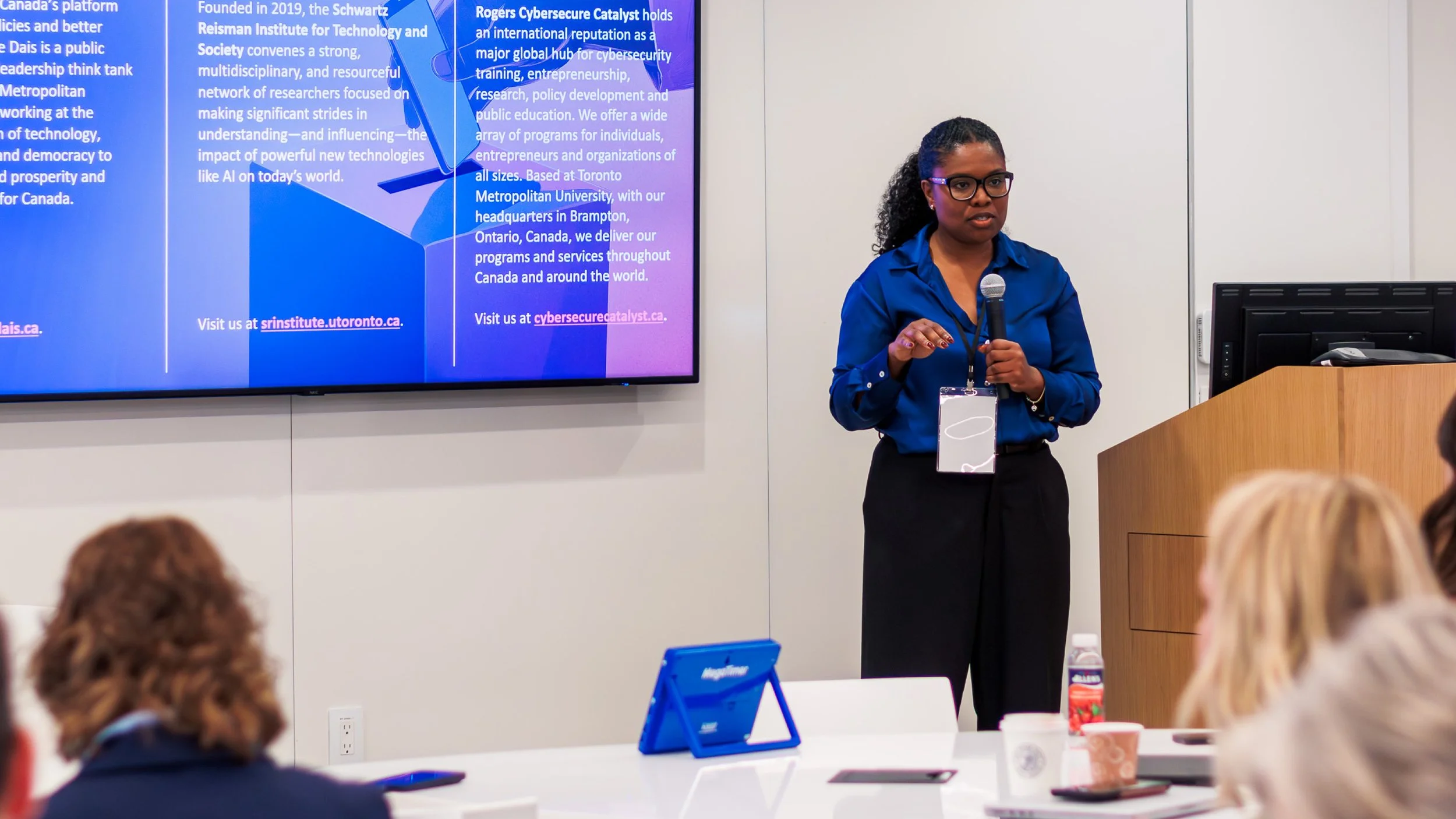 SRI Executive Director Monique Crichlow presents in front of a podium with a PowerPoint. She is wearing a dark blue shirt and holding a microphone.