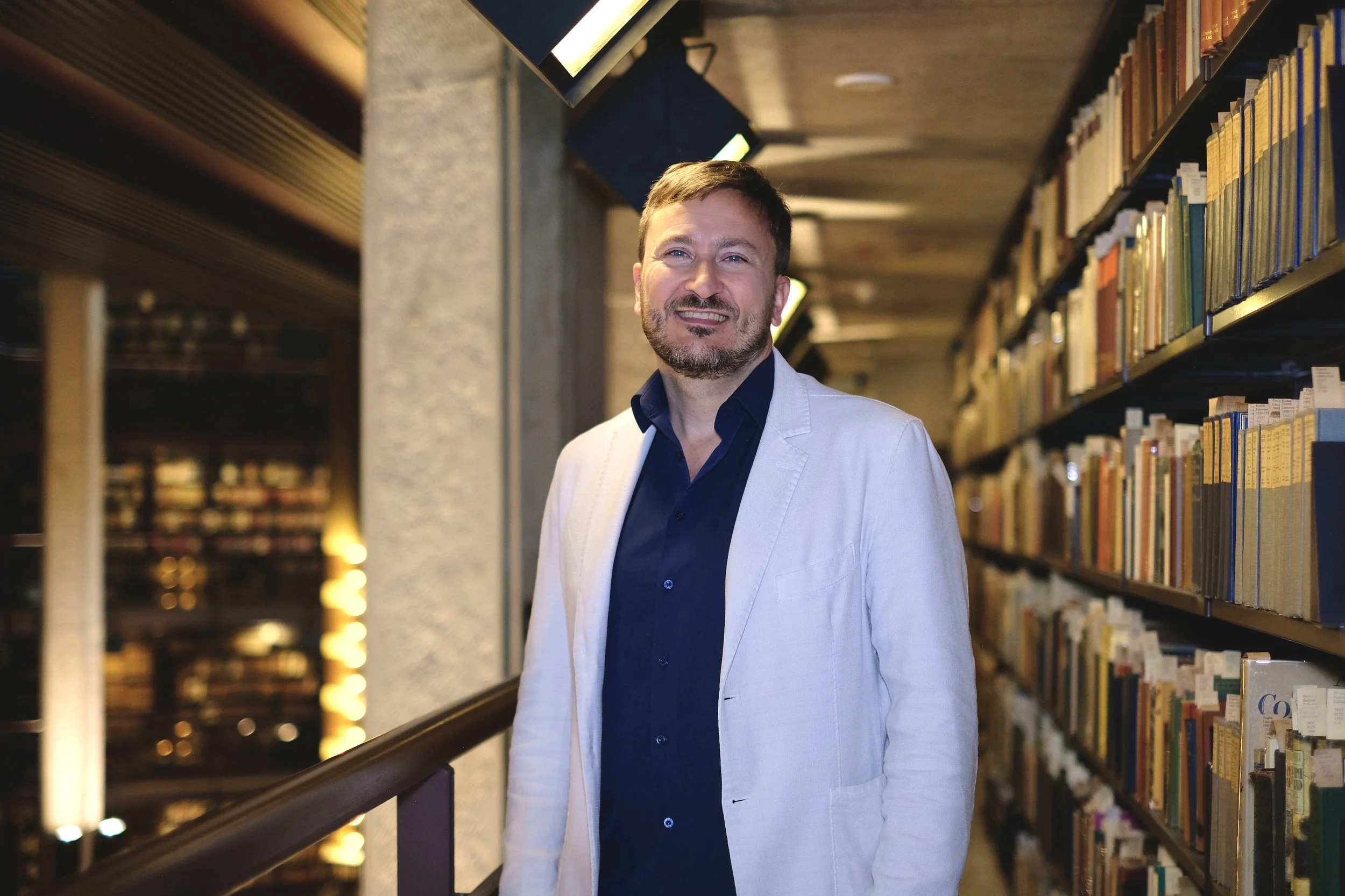 Paulo Granata stands wearing a white blazer and a blue shirt in the Fischer Rare Book Library at the University of Toronto.