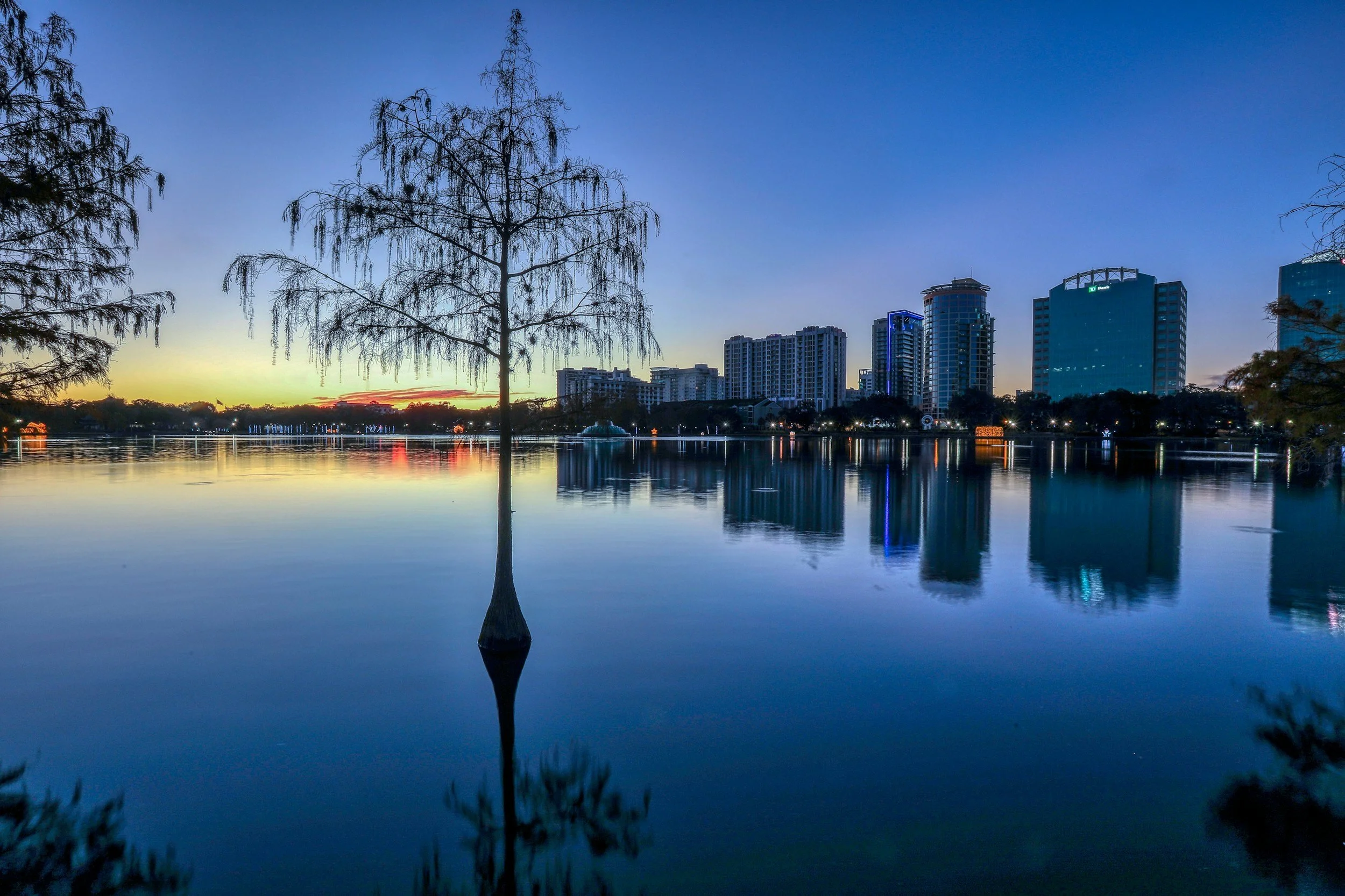 City skyline at sunset with buildings reflected on a calm body of water, trees in the foreground.
