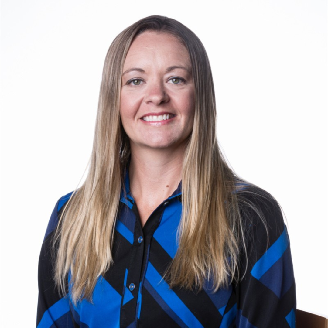 A woman with long blonde hair, wearing a blue and black patterned shirt, smiling at the camera against a plain white background.