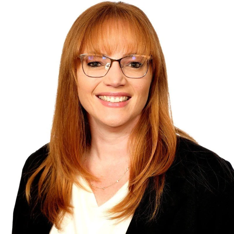 A woman with red hair, glasses, and a black blazer smiling. She is wearing a cream-colored top and a delicate gold necklace. The background is white.