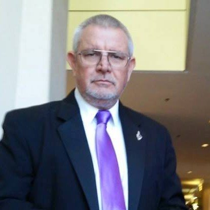 Man with gray hair and glasses wearing a black suit, white shirt, and purple tie, standing indoors with a light-colored wall and ceiling in the background.