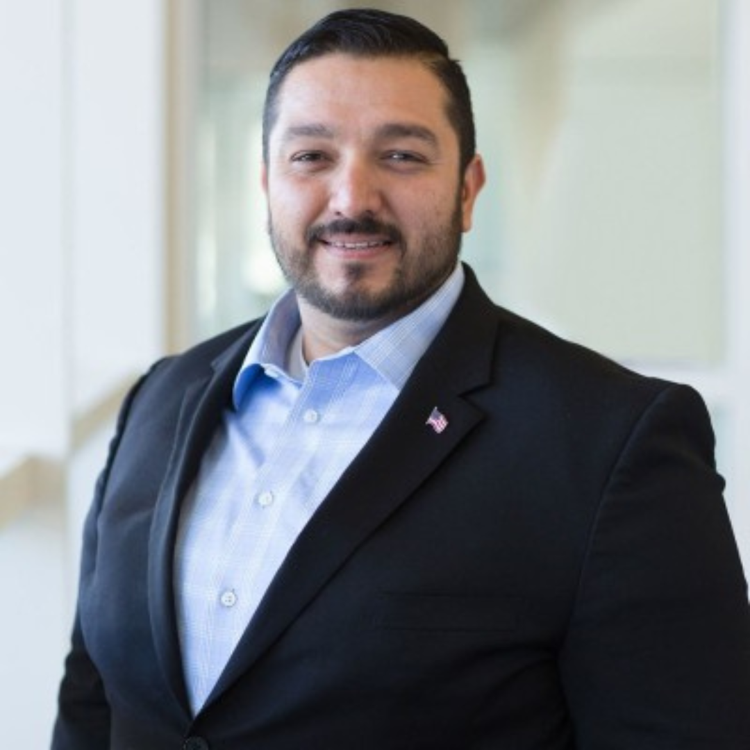 A man with dark hair and a beard wearing a dark blazer and a light blue collared shirt, smiling at the camera, with a small American flag pin on his blazer lapel.