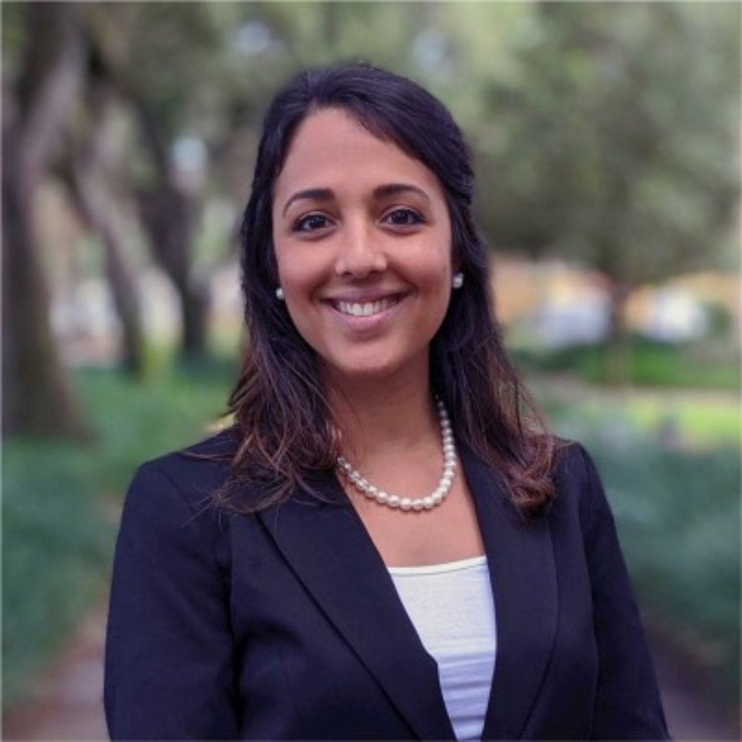 A woman with dark hair, wearing a black blazer, pearl necklace, and earrings, smiling outdoors with blurred trees in the background.