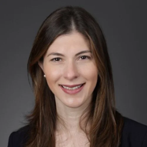 Headshot of a woman with long brown hair, smiling, wearing a black blazer against a plain gray background.