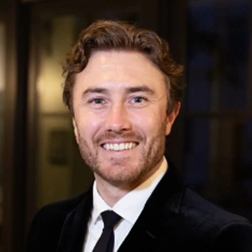 A man with curly brown hair and a beard, wearing a black suit and tie, smiling at the camera indoors.