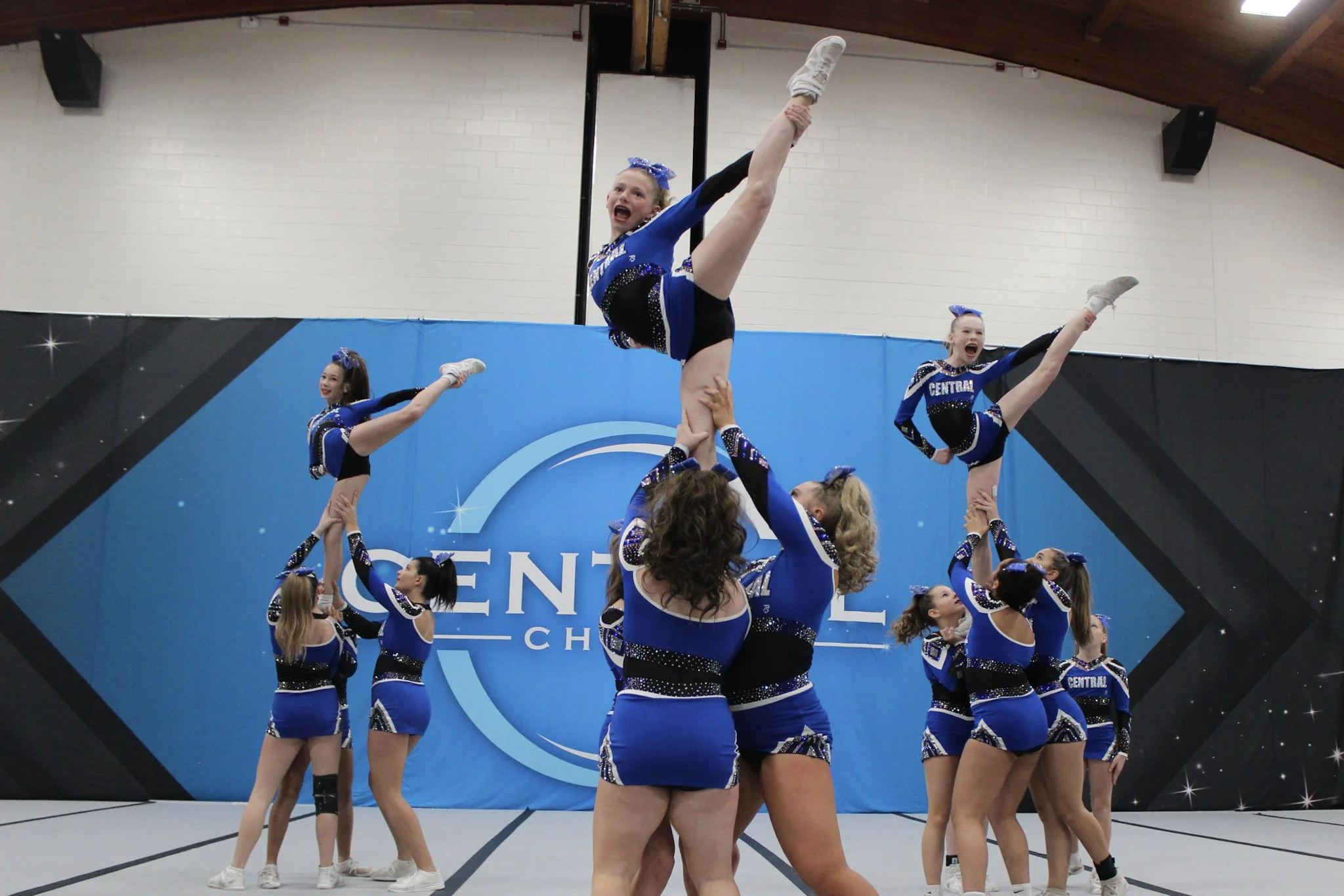A group of female gymnasts in pink uniforms with pink hair bows are practicing a lift, with one gymnast holding a pink ball overhead as they stand on a blue mat.
