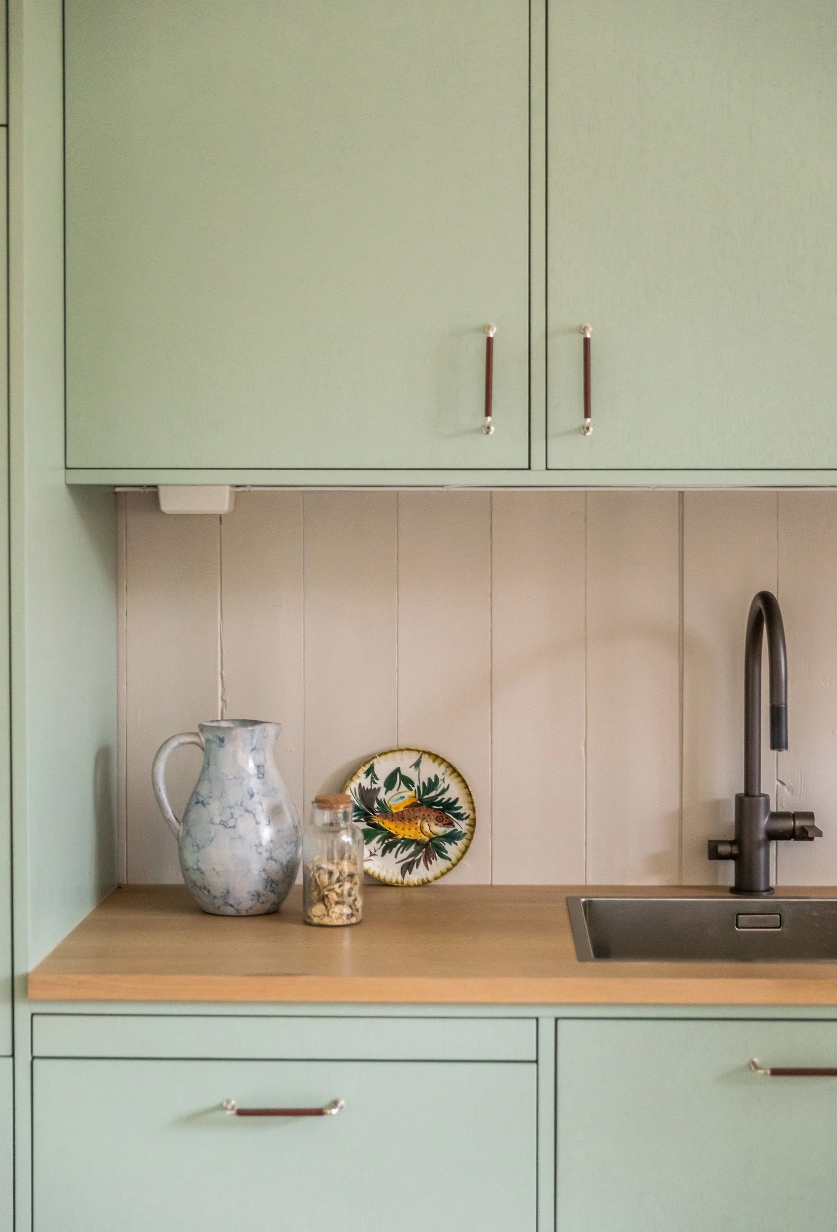 Detalj av grønt kjøkken med eikebenkeplate, lys flisvegg og mørk armatur.