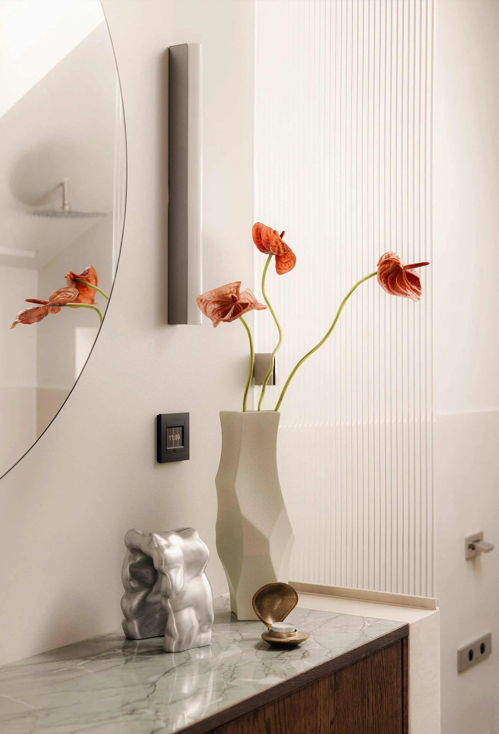 Detalj av servantinnredning med benkeplate i stein, stort speil og dekorative blomster mot rillet glassvegg.