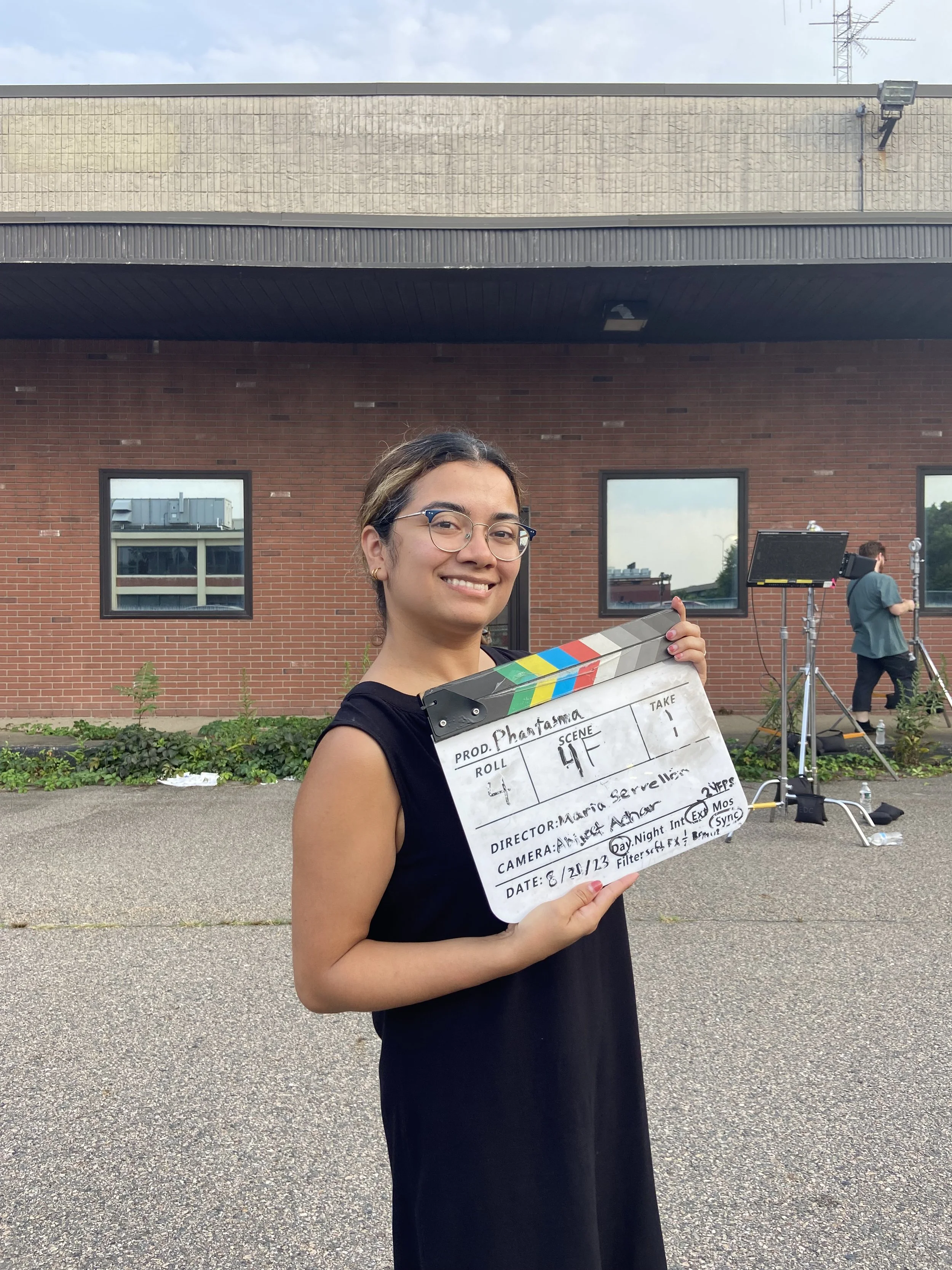 A young woman, Maria Servellon, in glasses holding a film clapboard on a film set outdoors, smiling at the camera.