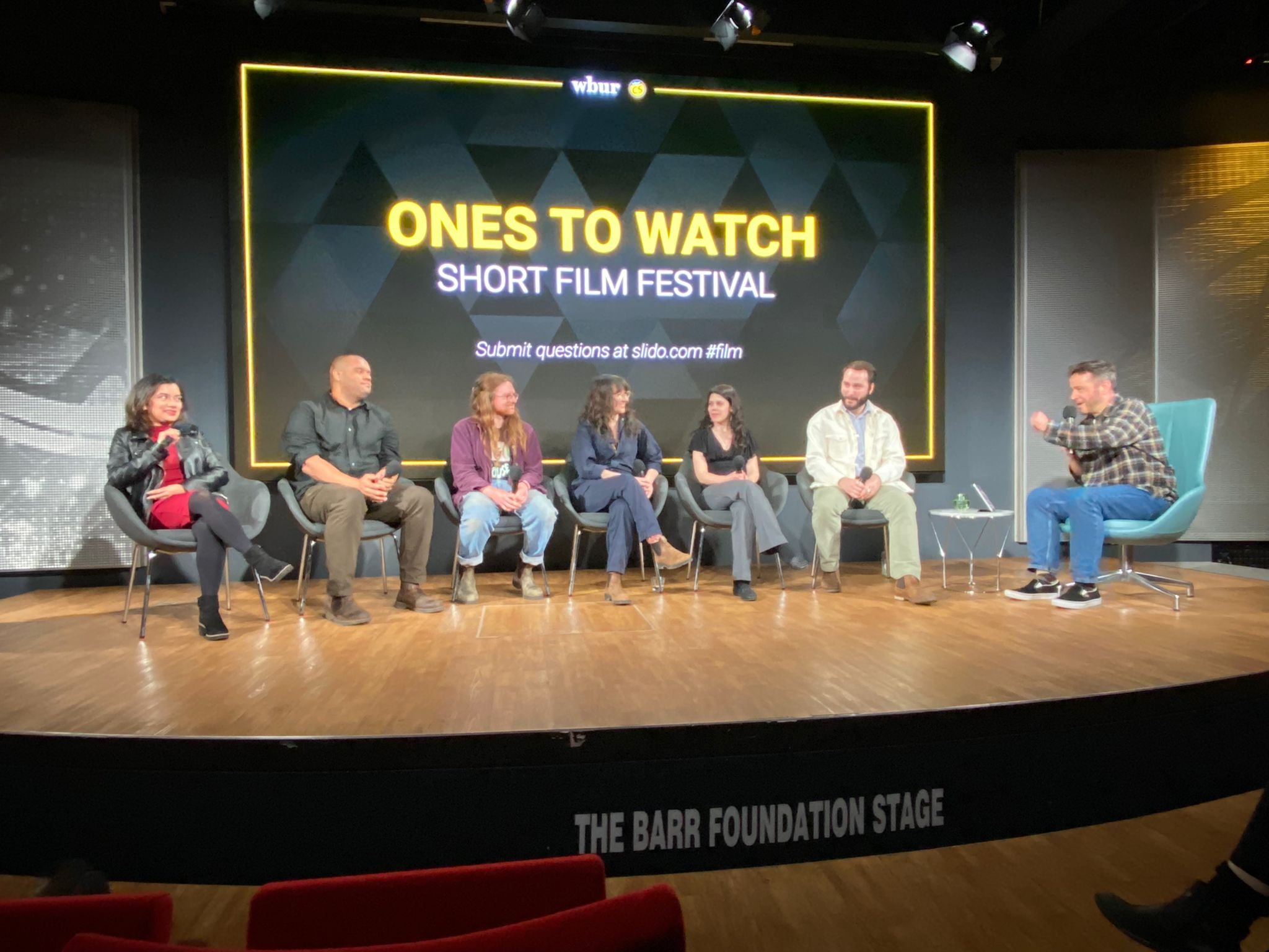 A panel discussion on a stage with seven people sitting, including Maria Servellon, during a short film festival event. The background screen reads 'ONES TO WATCH SHORT FILM FESTIVAL'.