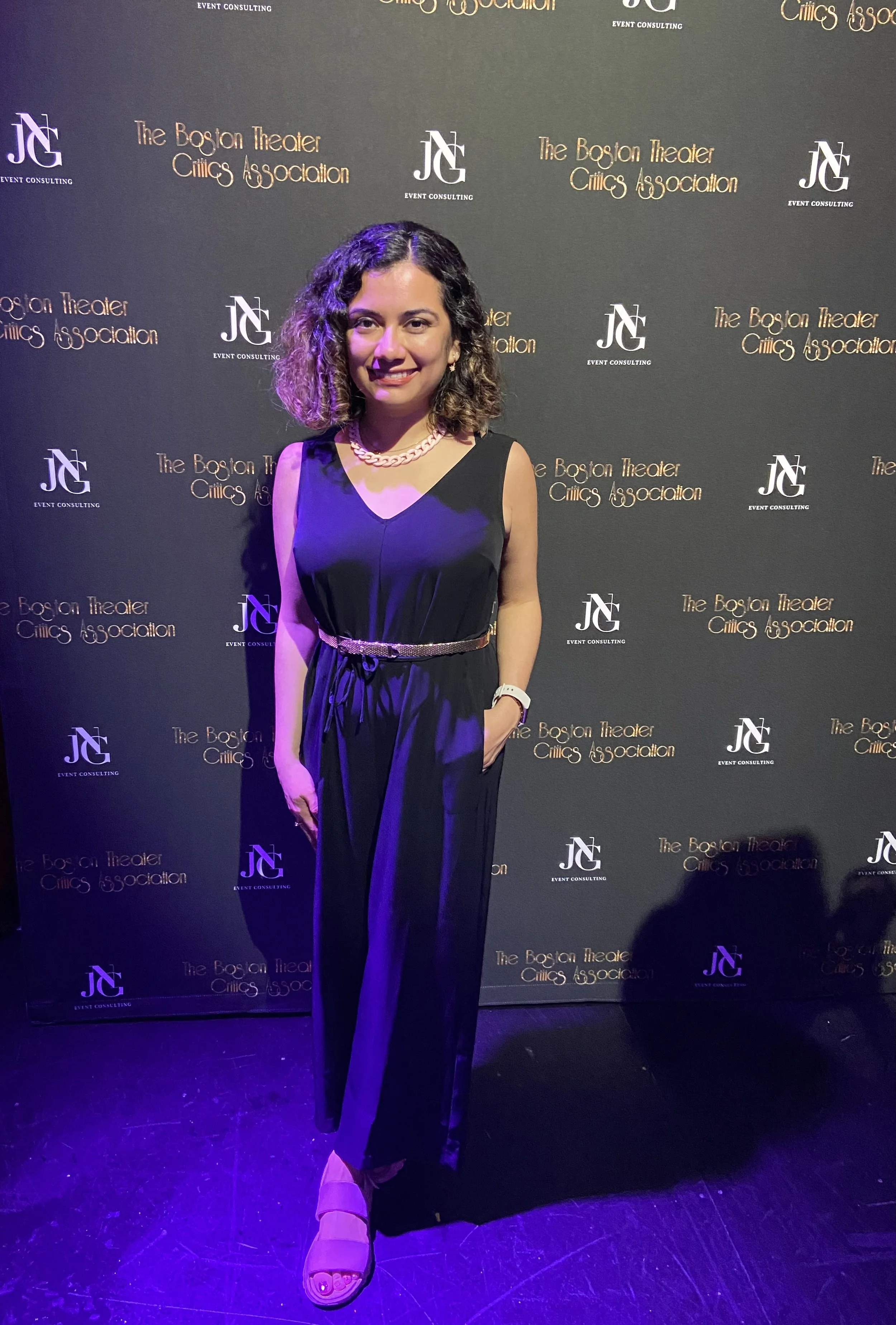 A woman, Maria Servellon, posing in a black sleeveless jumpsuit with a pearl necklace and a woven belt, standing in front of a step-and-repeat backdrop with logos for the Boston Theater Critics Association and JG Event Consulting, at an event.
