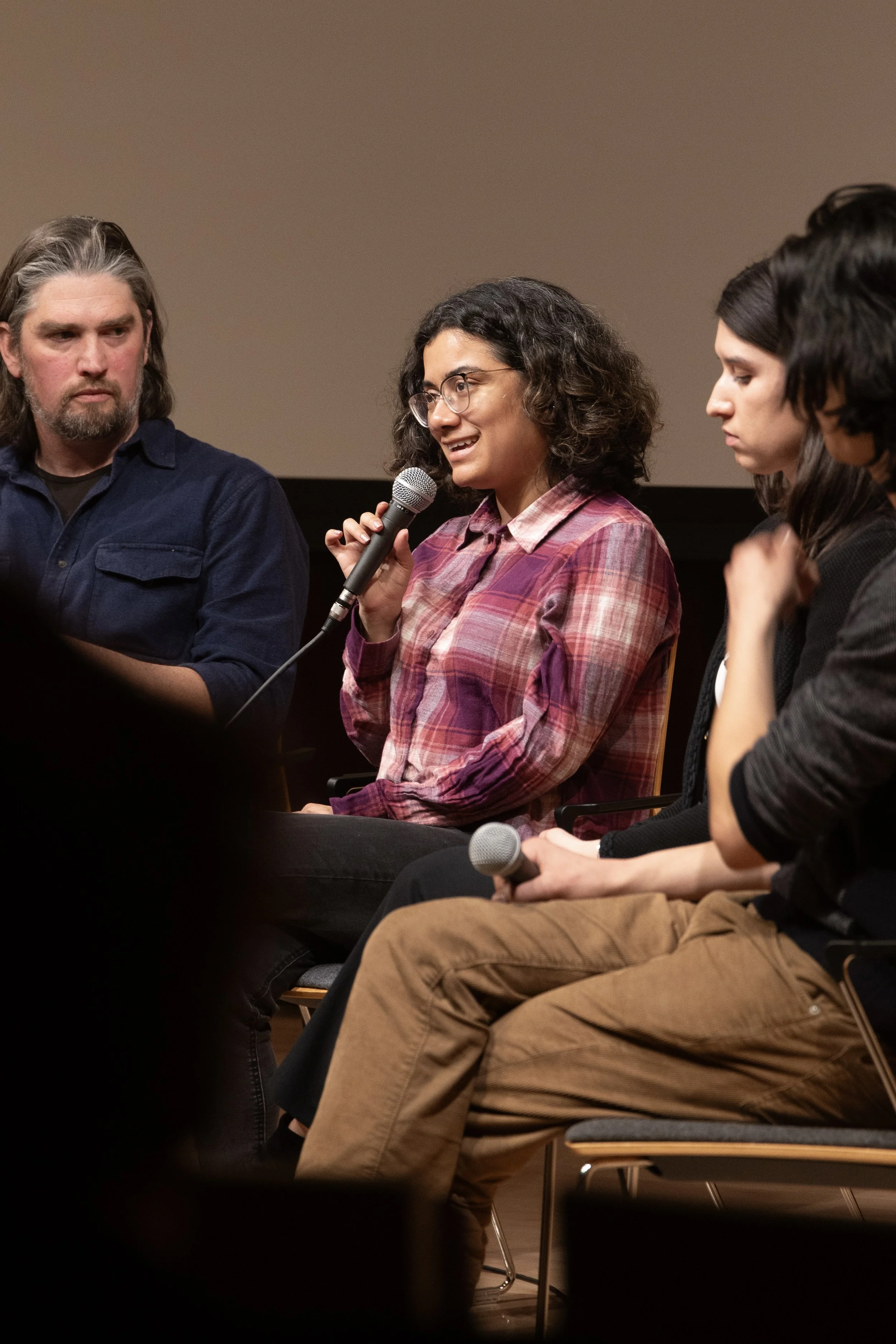 A woman, Maria Servellon, in a plaid shirt and glasses speaking into a microphone during a panel discussion, with two other men and one woman sitting beside her.